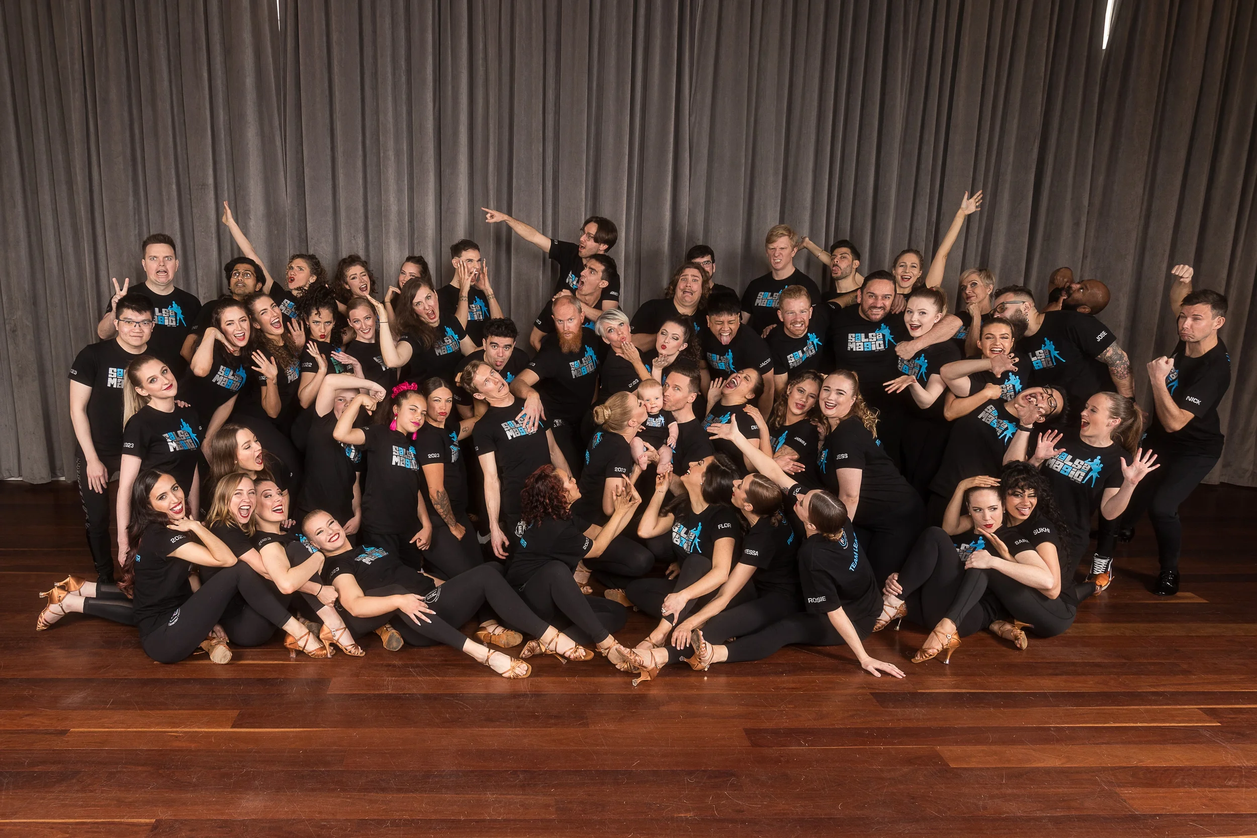 Group of people in black t-shirts on a wooden floor with gray curtains in the background, posing playfully for a group photo.