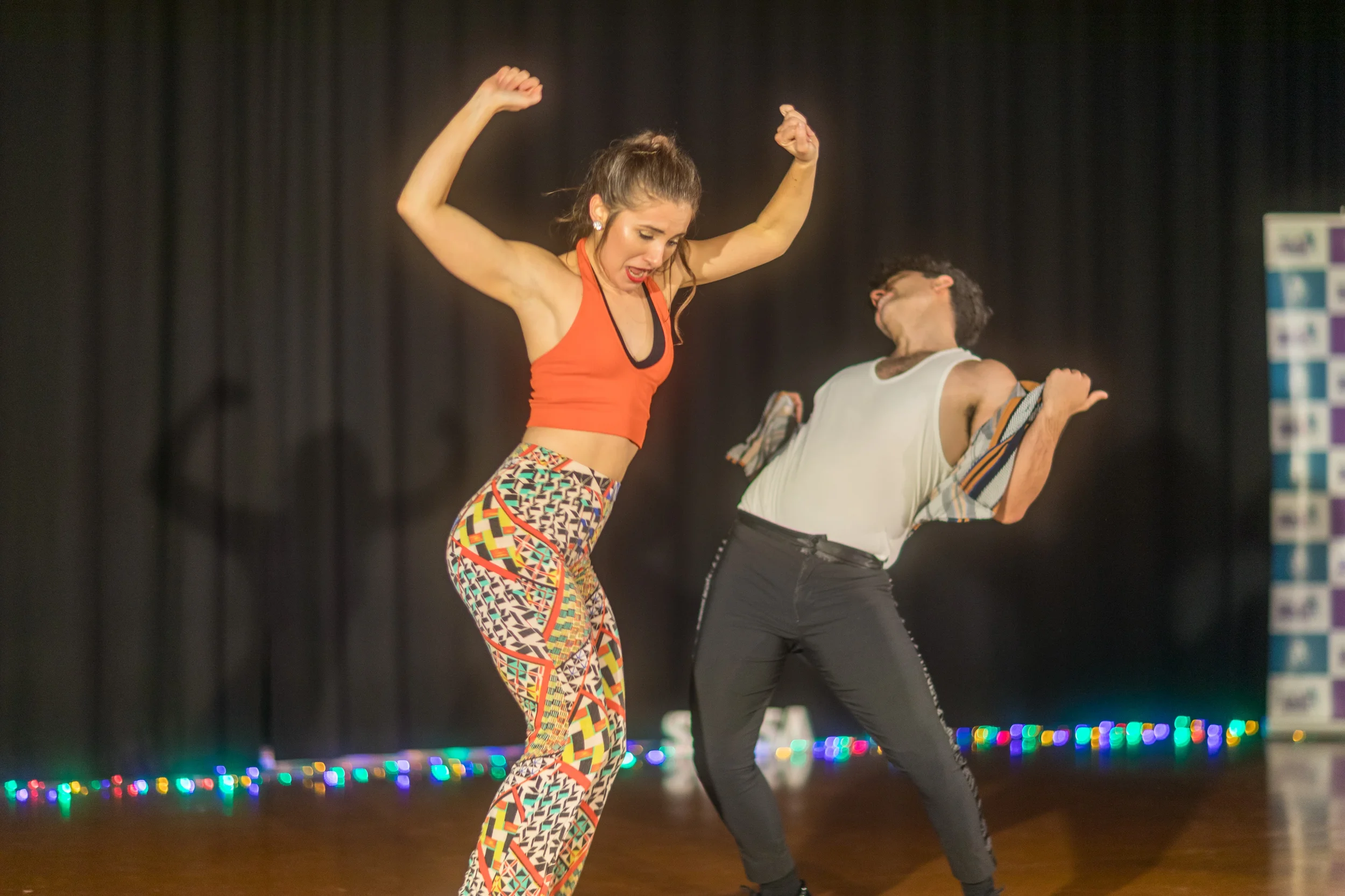 Two women are performing on stage with black curtains and colorful lights in the background, one is wearing a red crop top and patterned pants, the other in a white tank top and black pants.