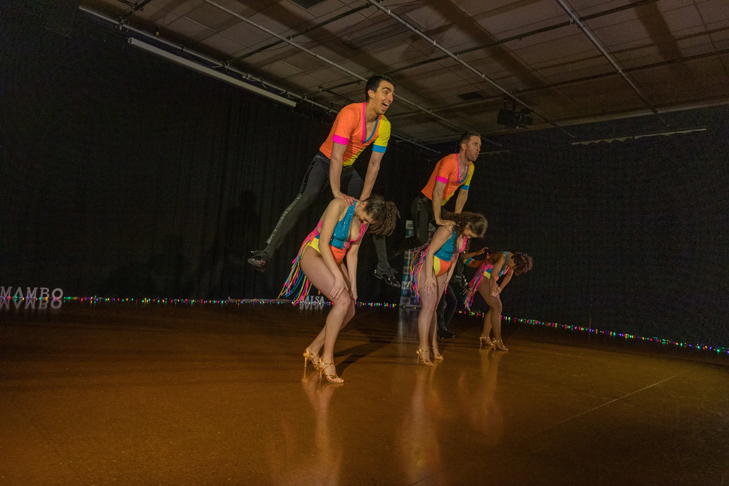 Six performers in colorful costumes performing a synchronized dance routine on stage, with three men on the top row and three women on the bottom, all striking a bent-over pose.