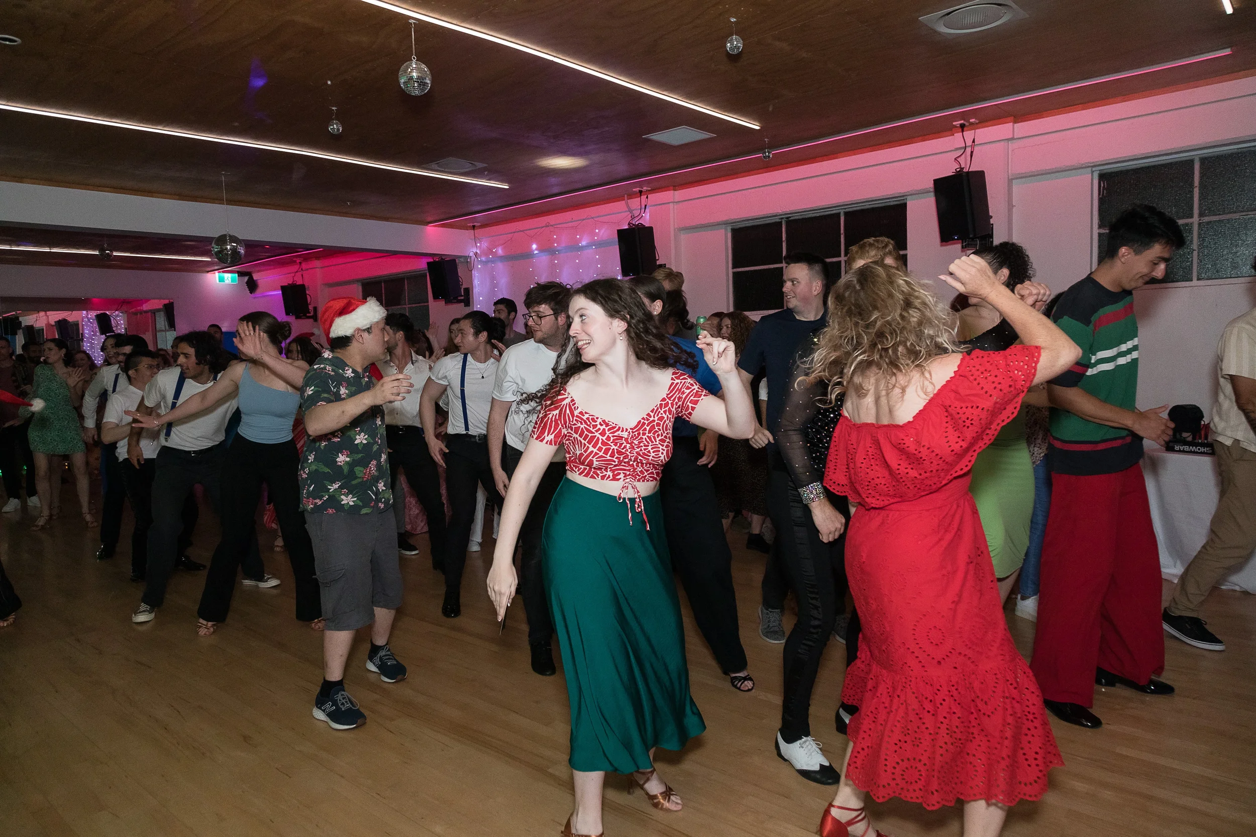People dancing at a party, some wearing festive or holiday attire, in a decorated indoor venue with pink and purple lighting.