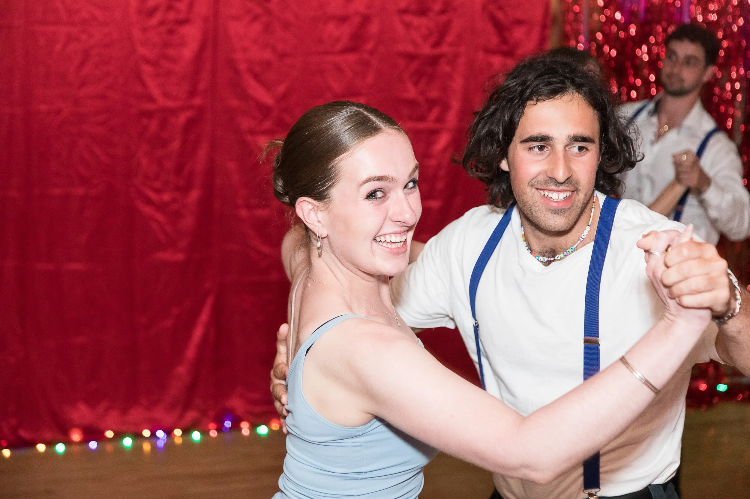 A man and a woman are dancing together, smiling, with a red, shiny curtain backdrop and colorful lights at the bottom.
