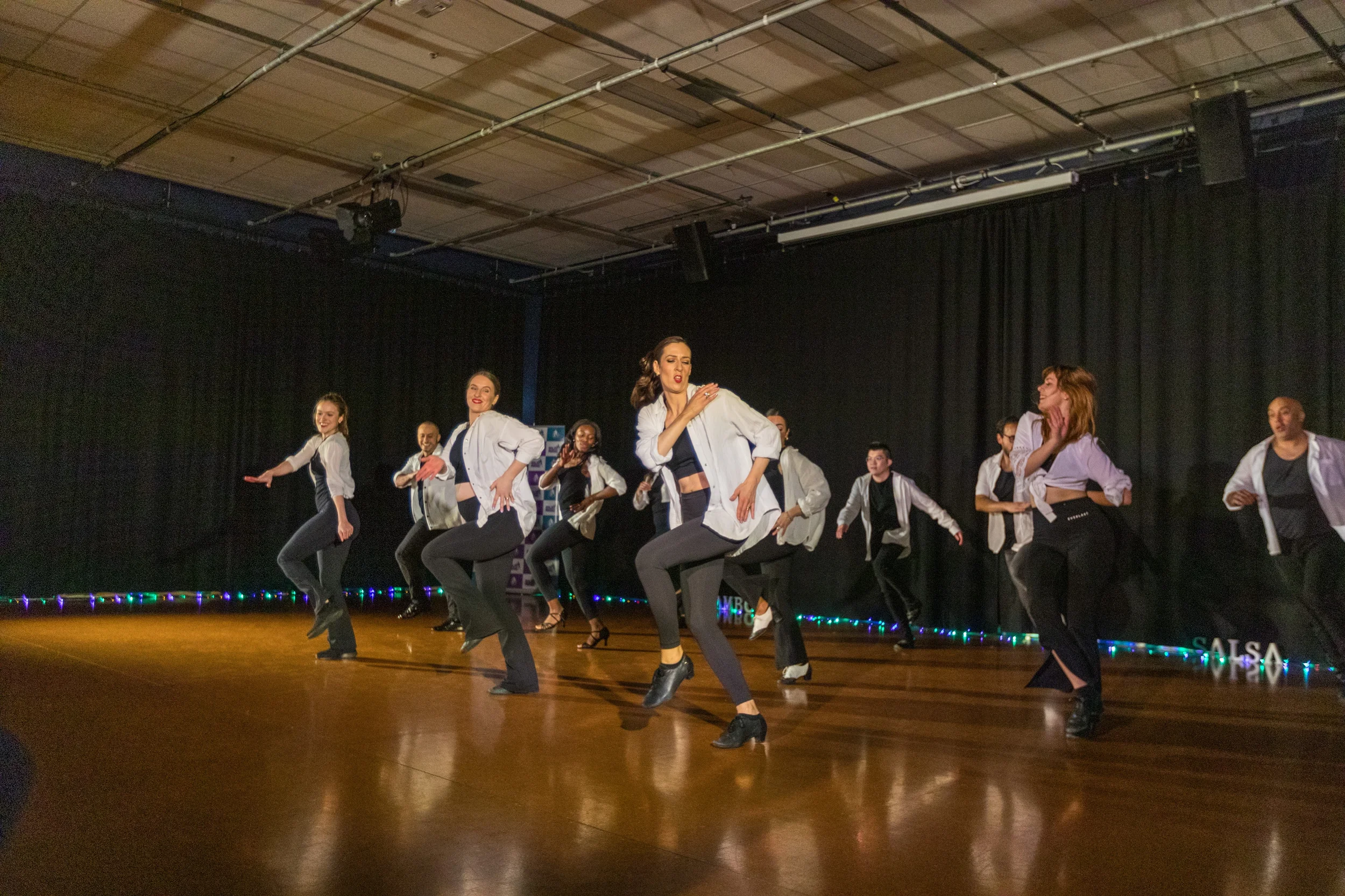 Group of dancers in white shirts and black pants performing on stage with black curtains and colorful lights.
