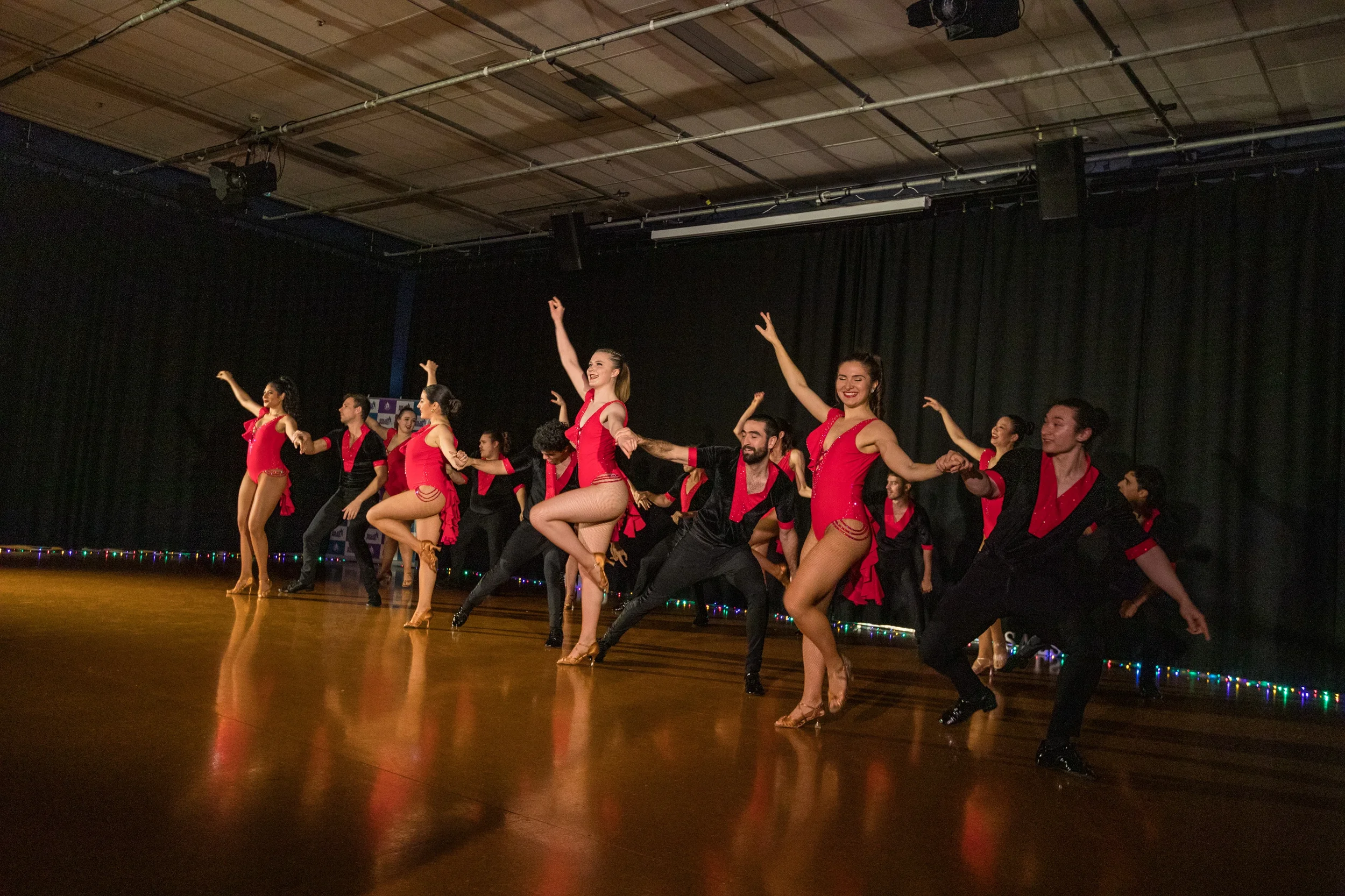 A group of dancers performing on stage with a black curtain backdrop, wearing costumes of red and black, mid-dance with varied poses, in a well-lit auditorium.