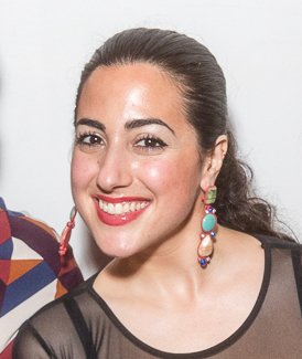 Close-up of a woman with dark hair pulled back, smiling, wearing large colorful earrings and a black top with a sheer neckline.