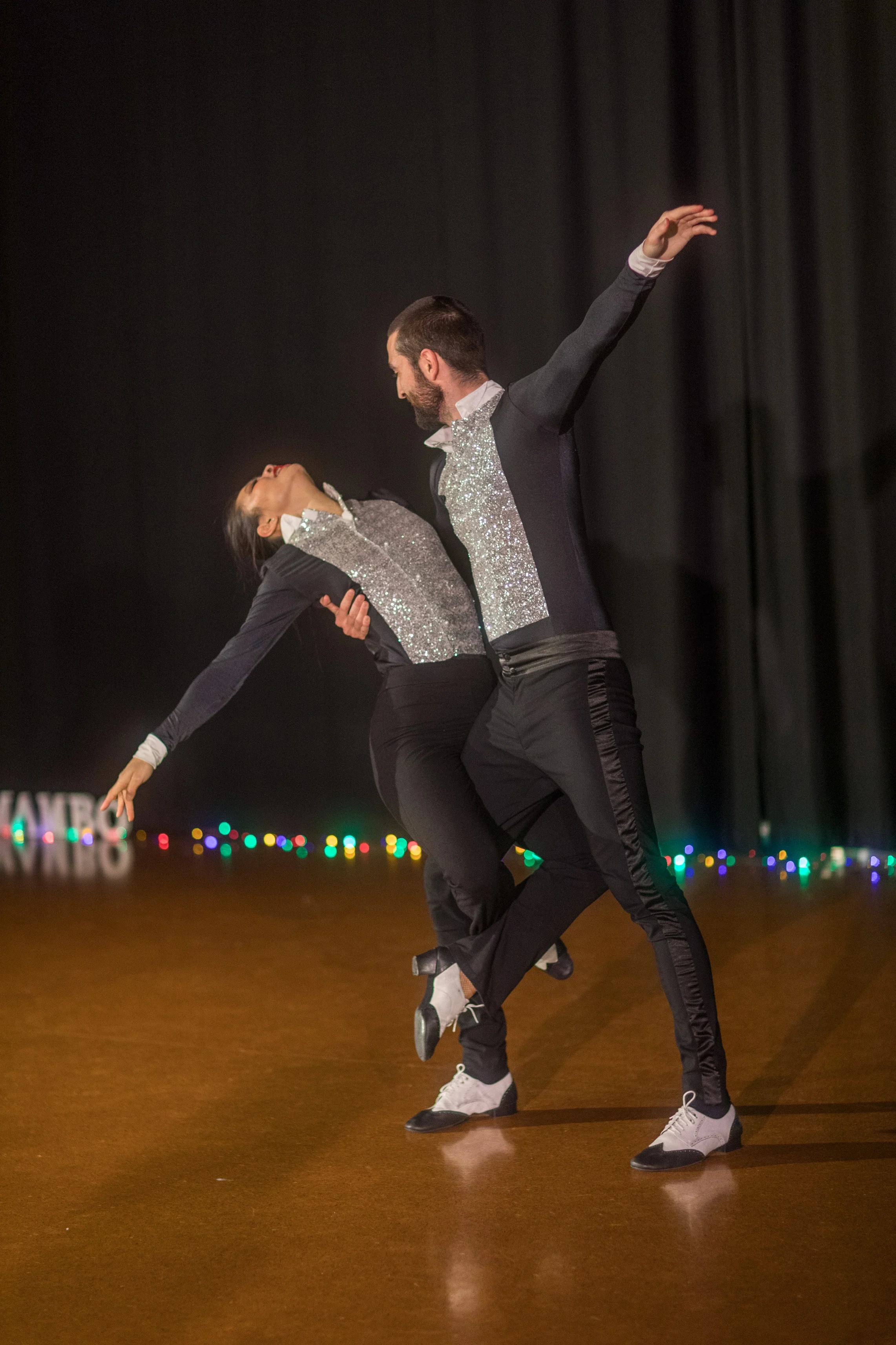 A man and woman engaged in a ballroom dance performance on a stage with black curtains and colorful lights in the background.