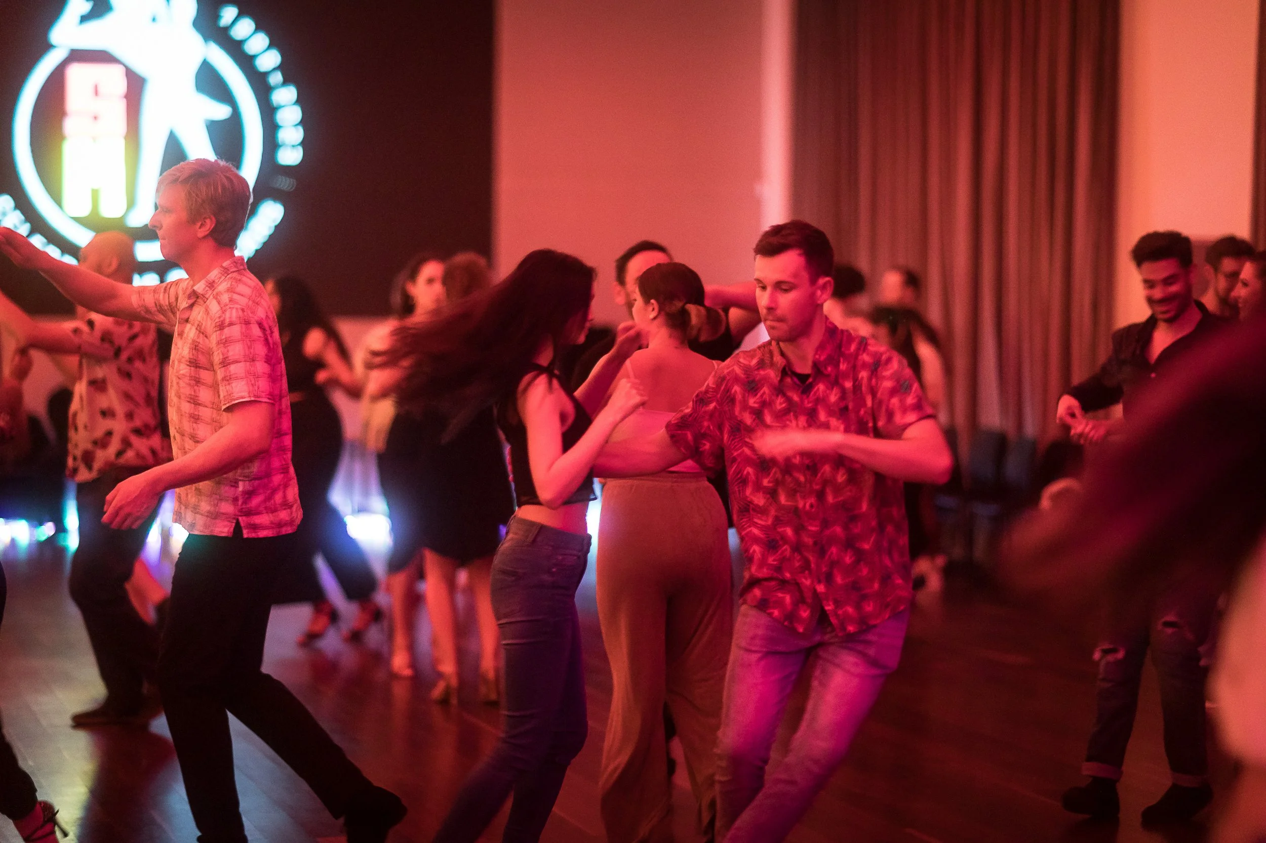 People dancing in a dimly lit room with a large logo on a screen in the background.