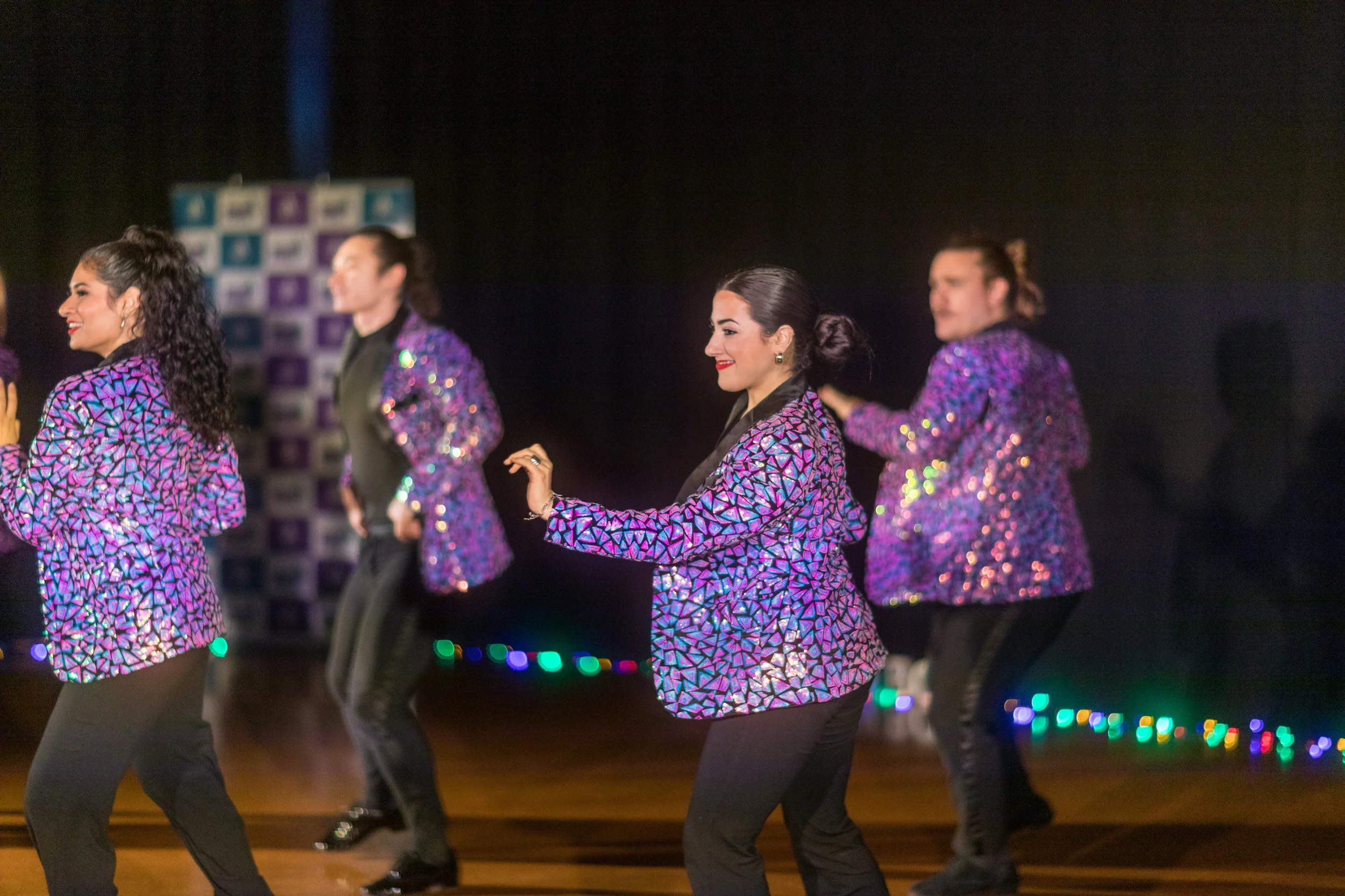 Four women performing a dance on stage, wearing matching purple sequined jackets and black pants.