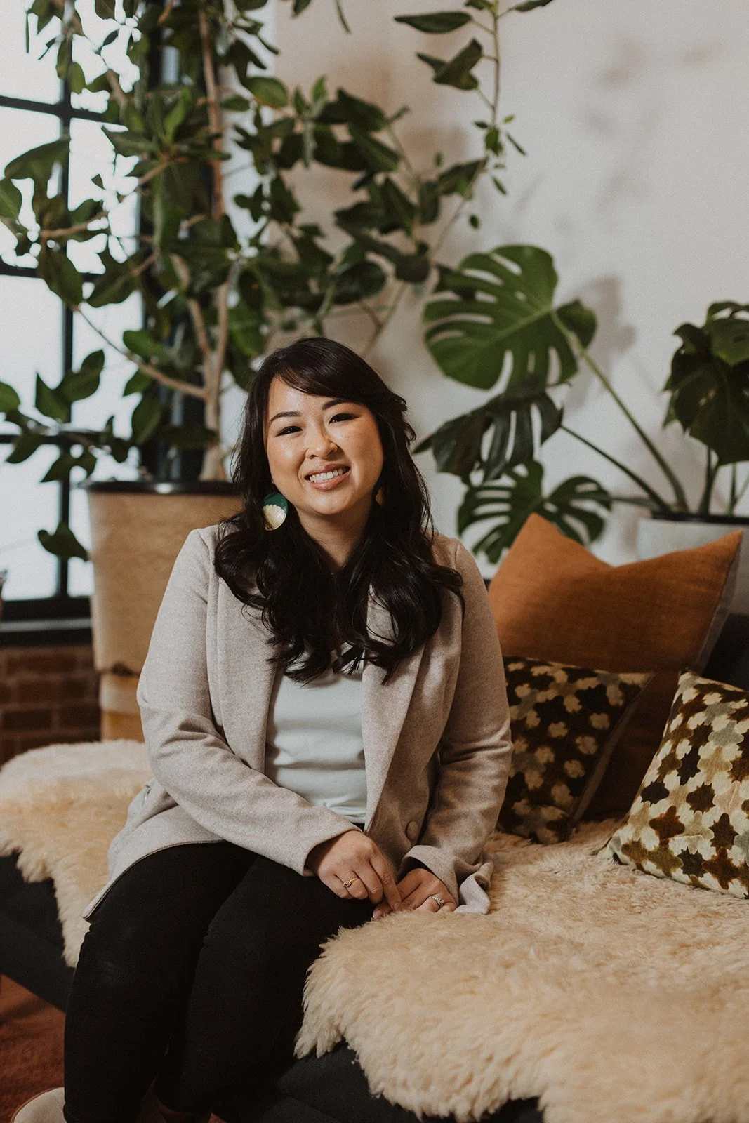 A woman sitting on a black sofa with beige and patterned pillows, smiling and looking at the camera, with a large leafy plant and abstract art on the wall behind her.