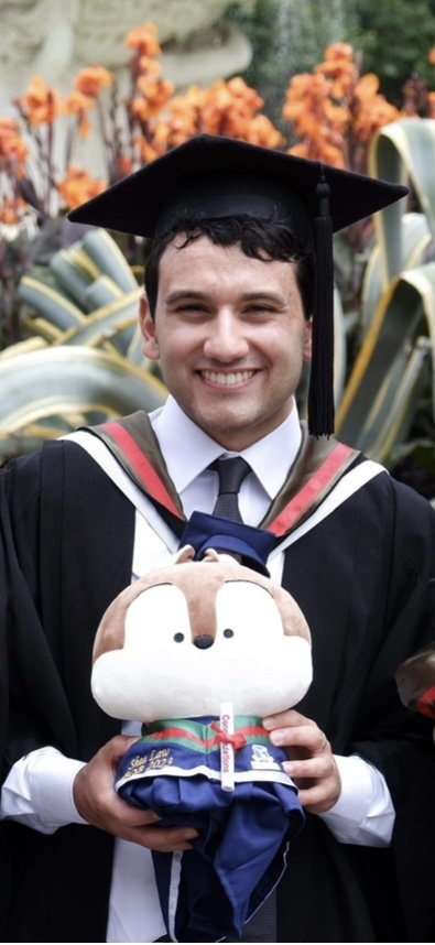A young wedding violinist in a graduation cap and gown holding a stuffed animal in front of a garden with orange flowers.