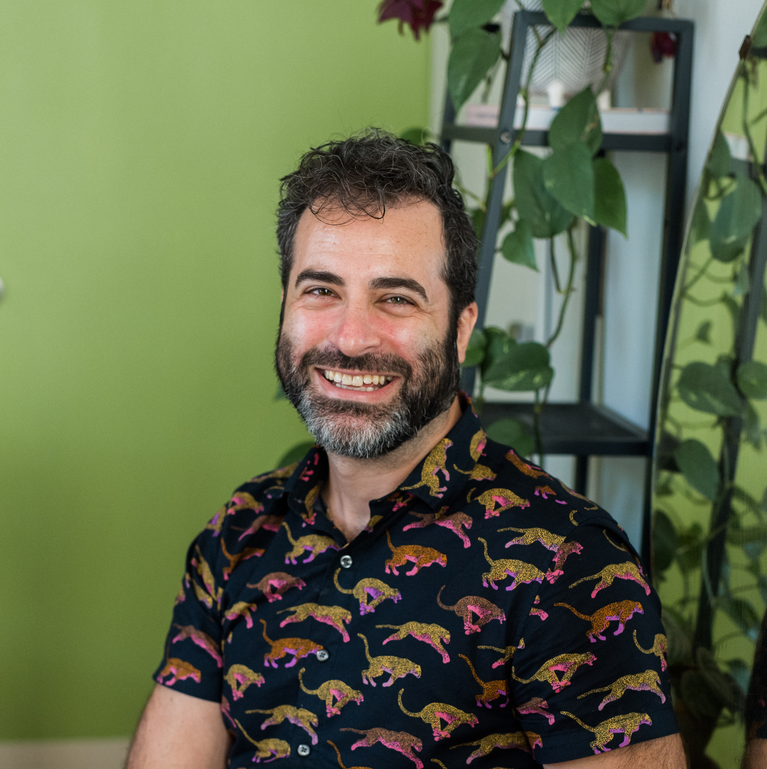 Close-up of a smiling man with dark, curly hair and a beard, wearing a black shirt with colorful dinosaur patterns, standing in front of a green wall and a black shelving unit with green plant leaves.