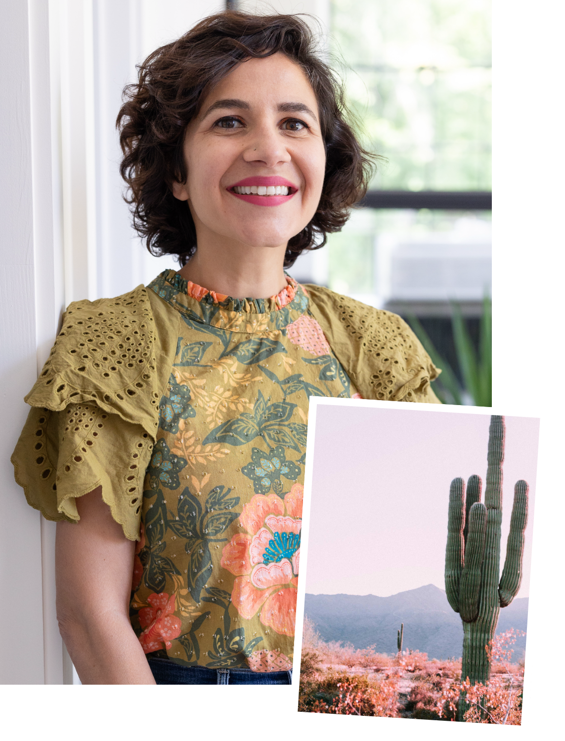 A smiling woman with short curly brown hair, wearing a floral patterned top with yellow lace sleeves, standing in front of a window, holding an image of a desert landscape with a large saguaro cactus and mountains in the background.
