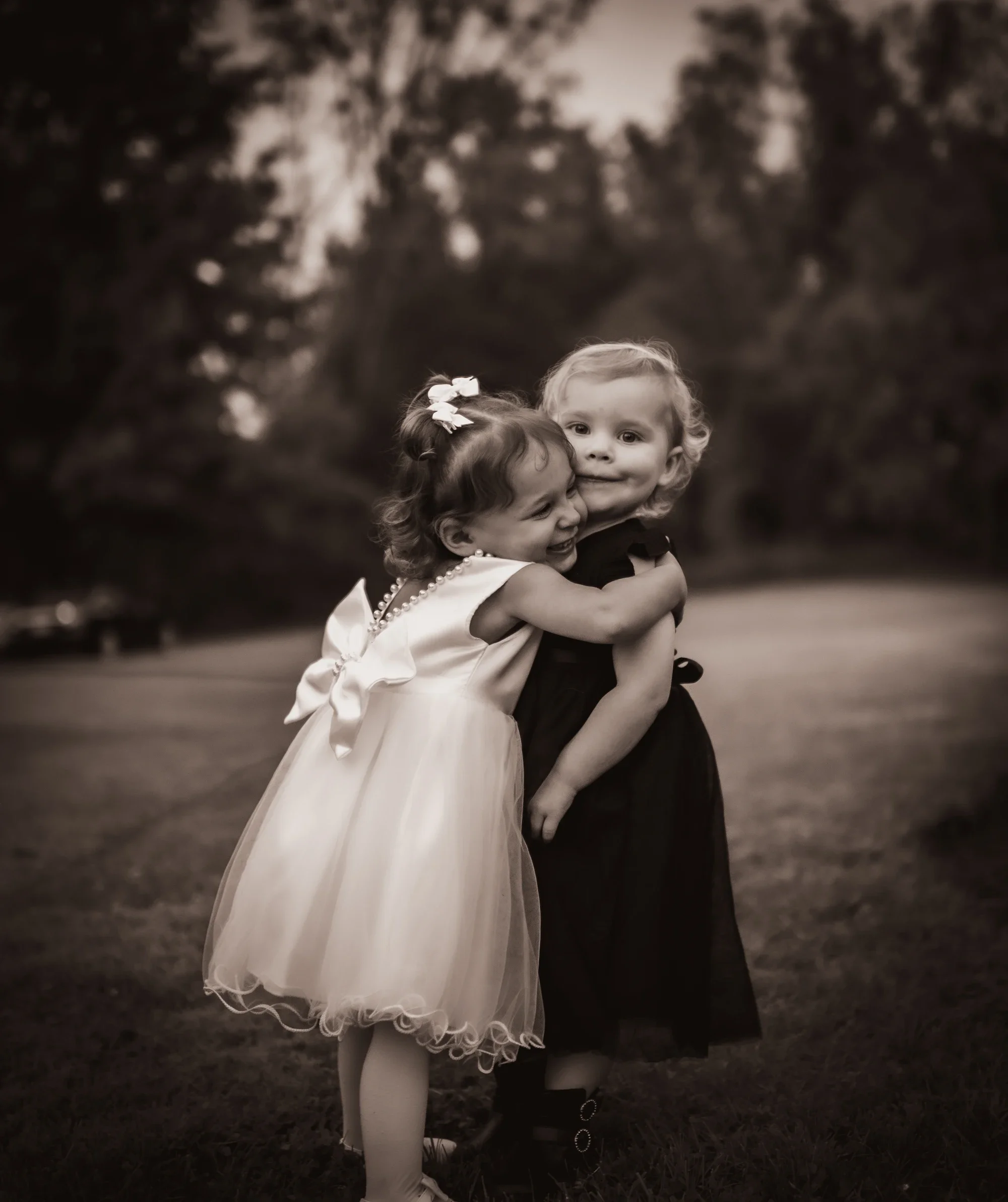 Two young girls, dressed in fancy dresses, happily hugging each other outdoors in a park, with trees in the background.