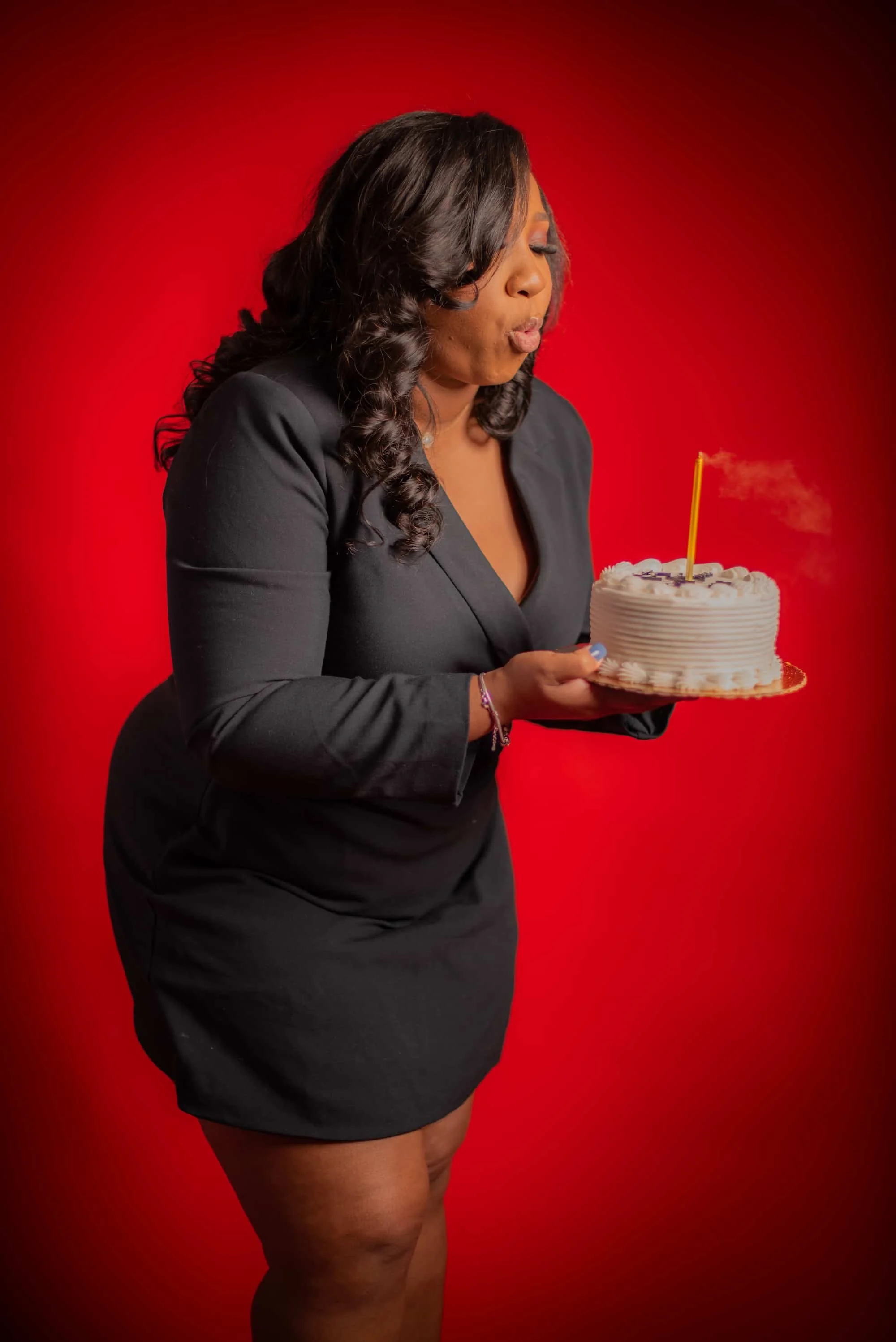 A woman in a black dress holding a birthday cake with a yellow candle against a red background.