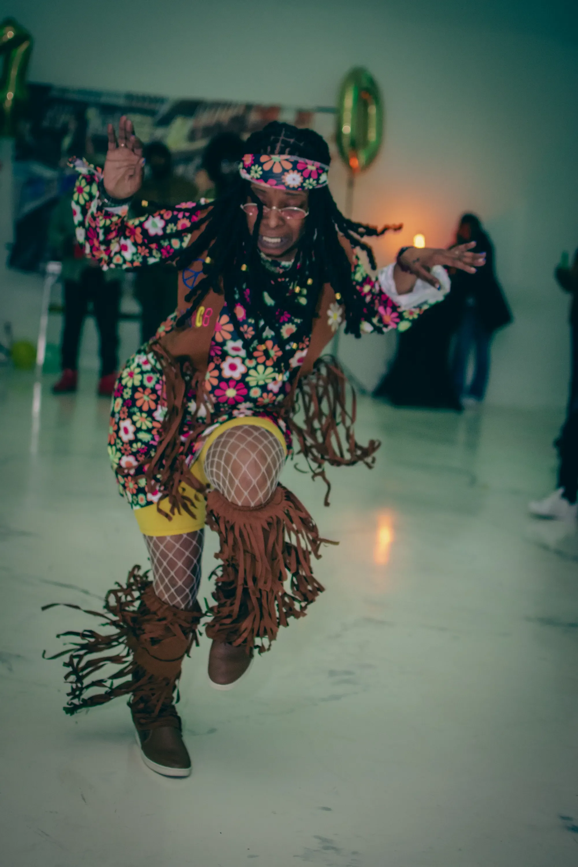 A woman dressed in a colorful 80's outfit with fringe and fishnet stockings is dancing energetically in a room with other people in the background. She has long long hair, wearing glasses and a headband, and appears joyful.
