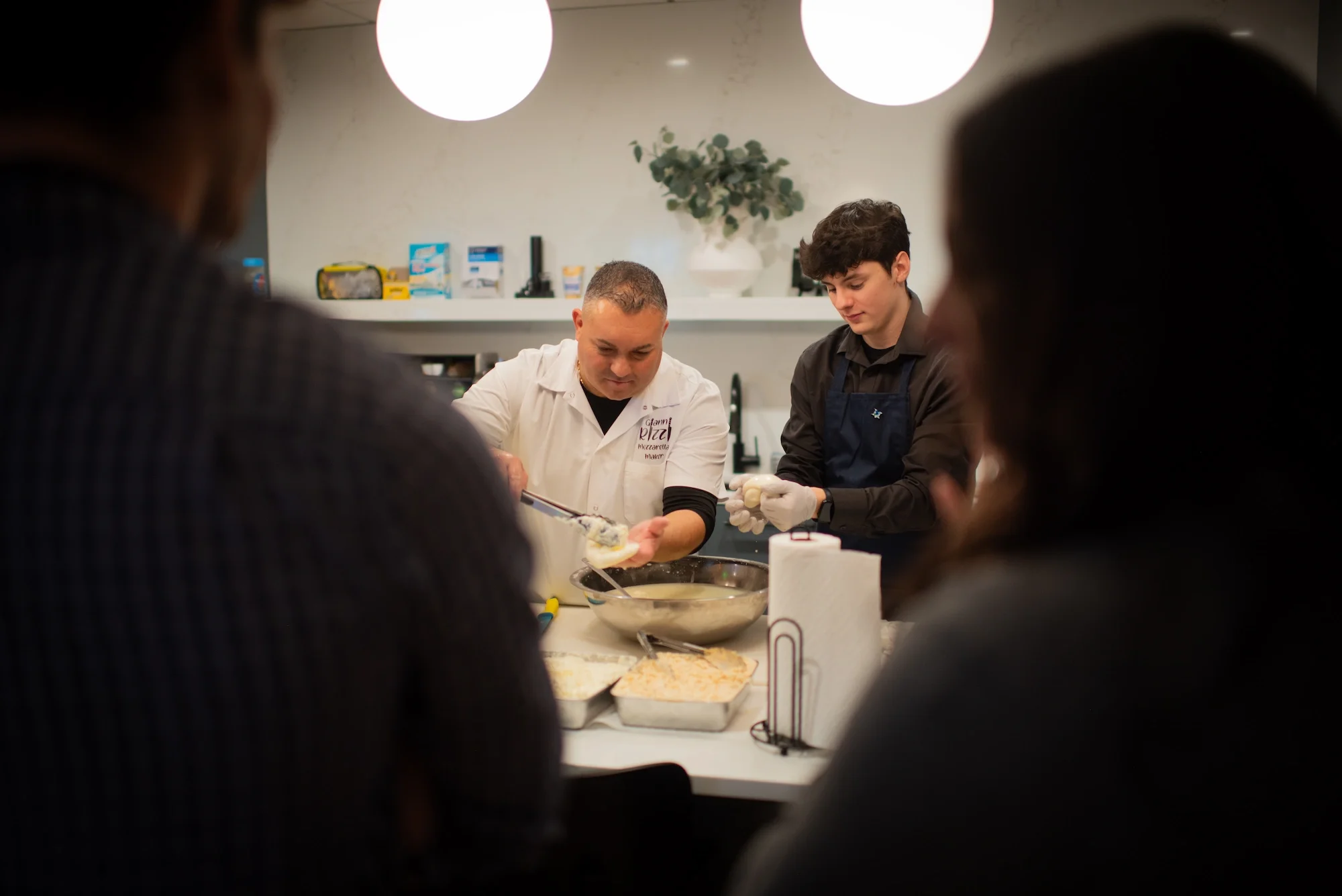 Two chefs preparing food in a kitchen while a group of people observe from in front of the camera.