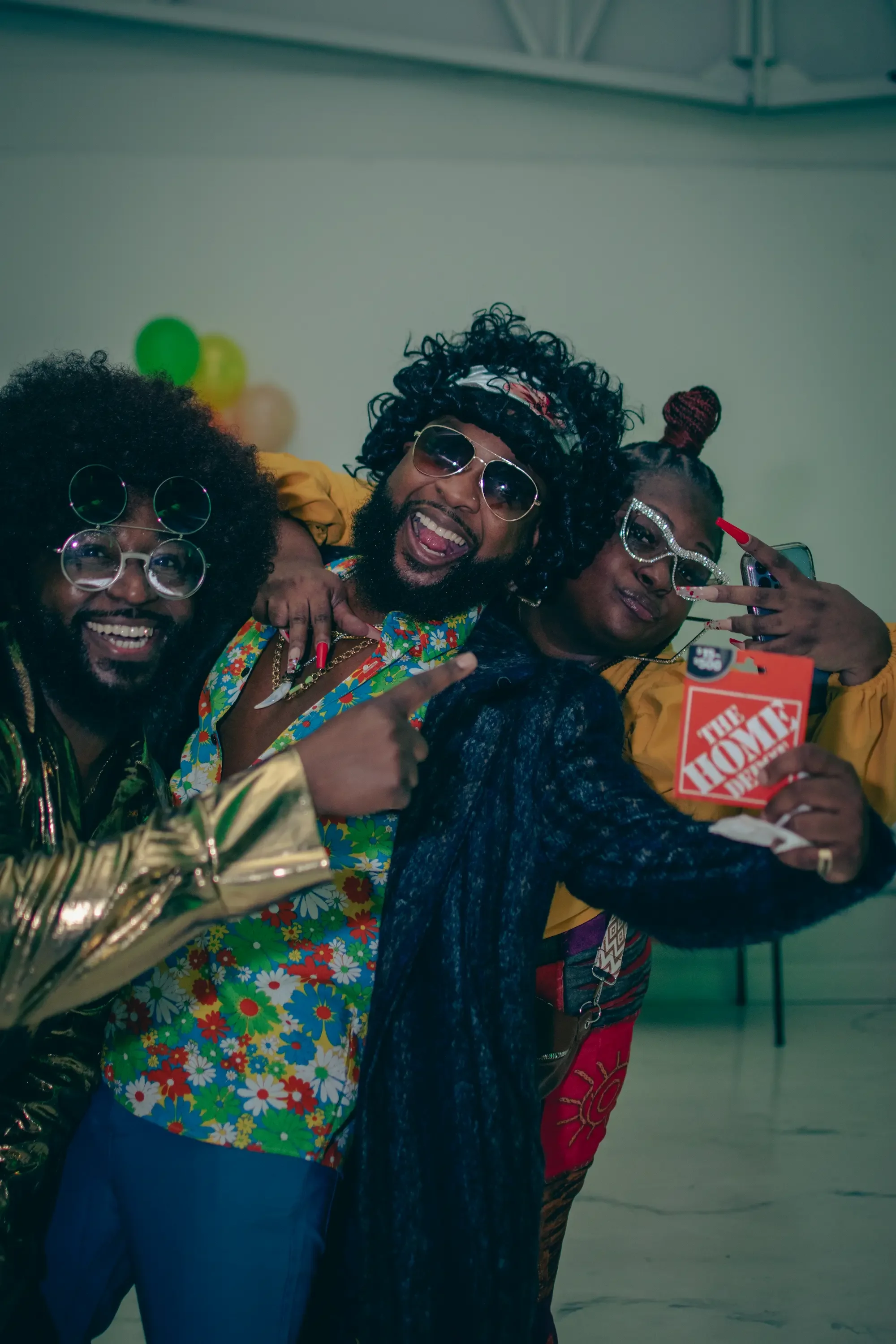 Three people dressed in colorful, retro 70s costumes taking a photo together indoors with balloons in the background.
