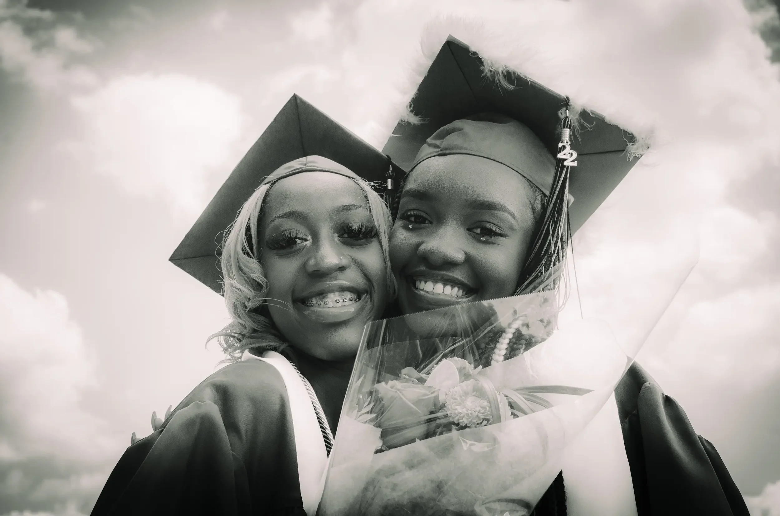 Two young women in graduation caps and gowns smiling, one holding a bouquet of flowers, against a cloudy sky background.