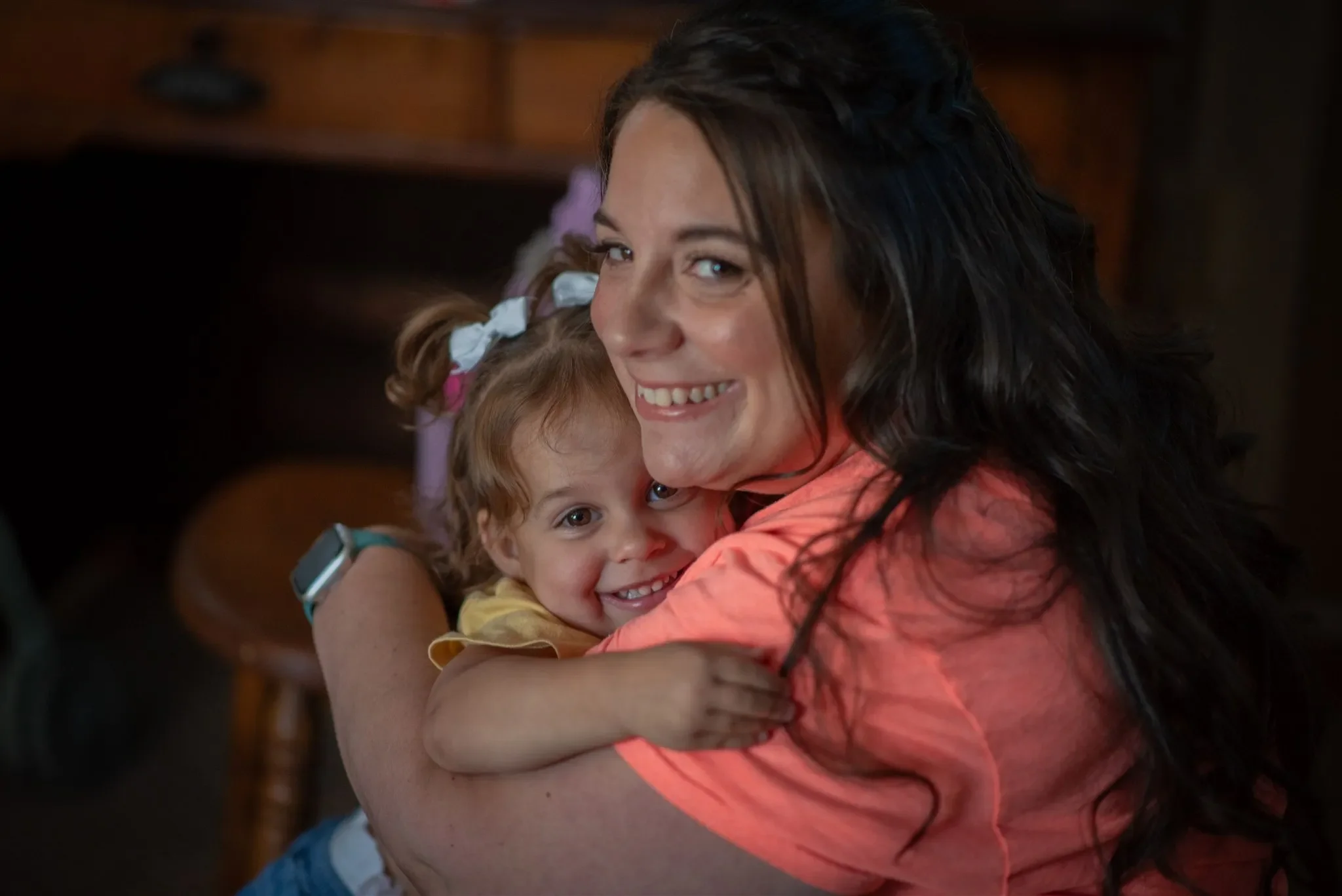 A woman and a young girl hugging each other indoors. The woman has dark wavy hair and is smiling into the camera. The girl has curly hair with bows, smiling happily. They appear to be enjoying a warm, affectionate moment.
