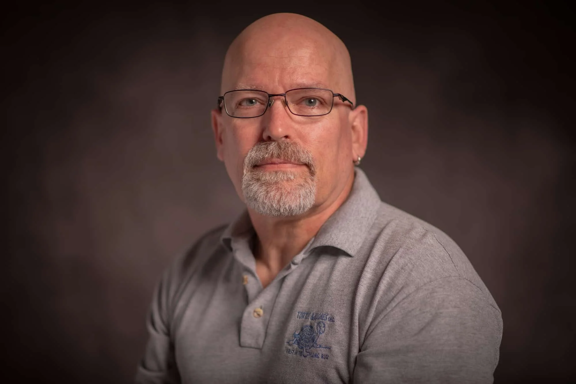 A middle-aged man with glasses and a beard, wearing a gray polo shirt, posing against a dark background.