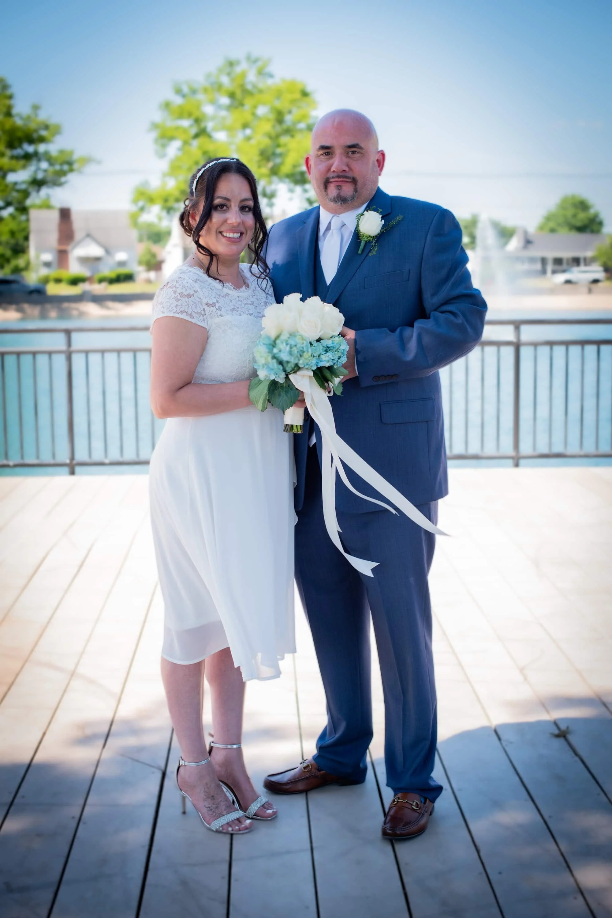 A bride and groom standing on a wooden deck near a body of water, smiling for a photo on their wedding day, with trees and houses in the background.