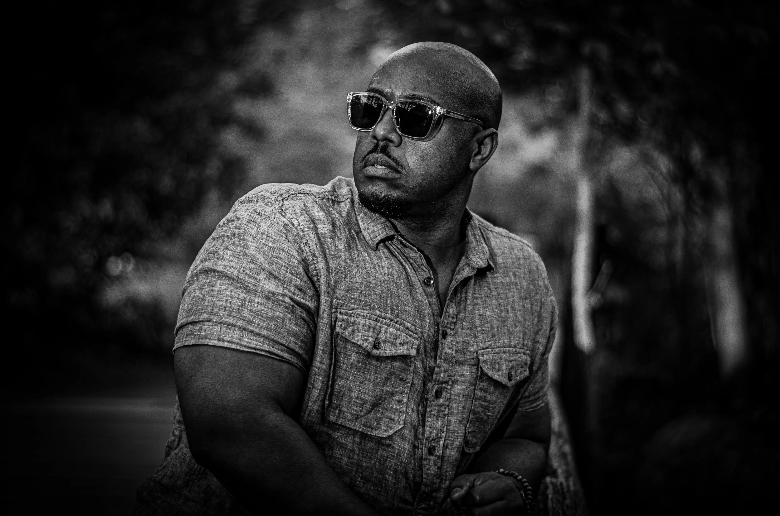 A man with dark sunglasses and a patterned short-sleeve shirt looking to the side outdoors with a blurred natural background in black and white.
