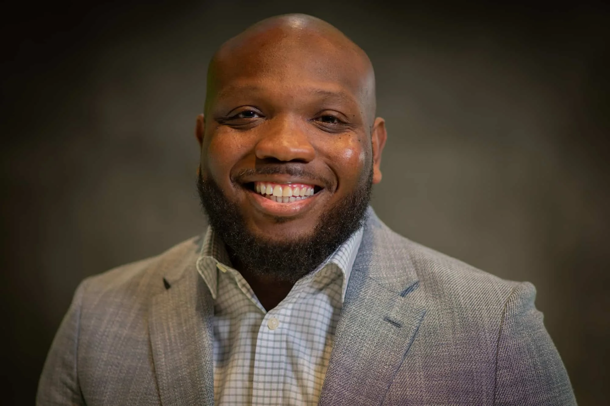 A smiling Black man with a beard, wearing a light gray blazer and a checkered shirt, against a dark background.
