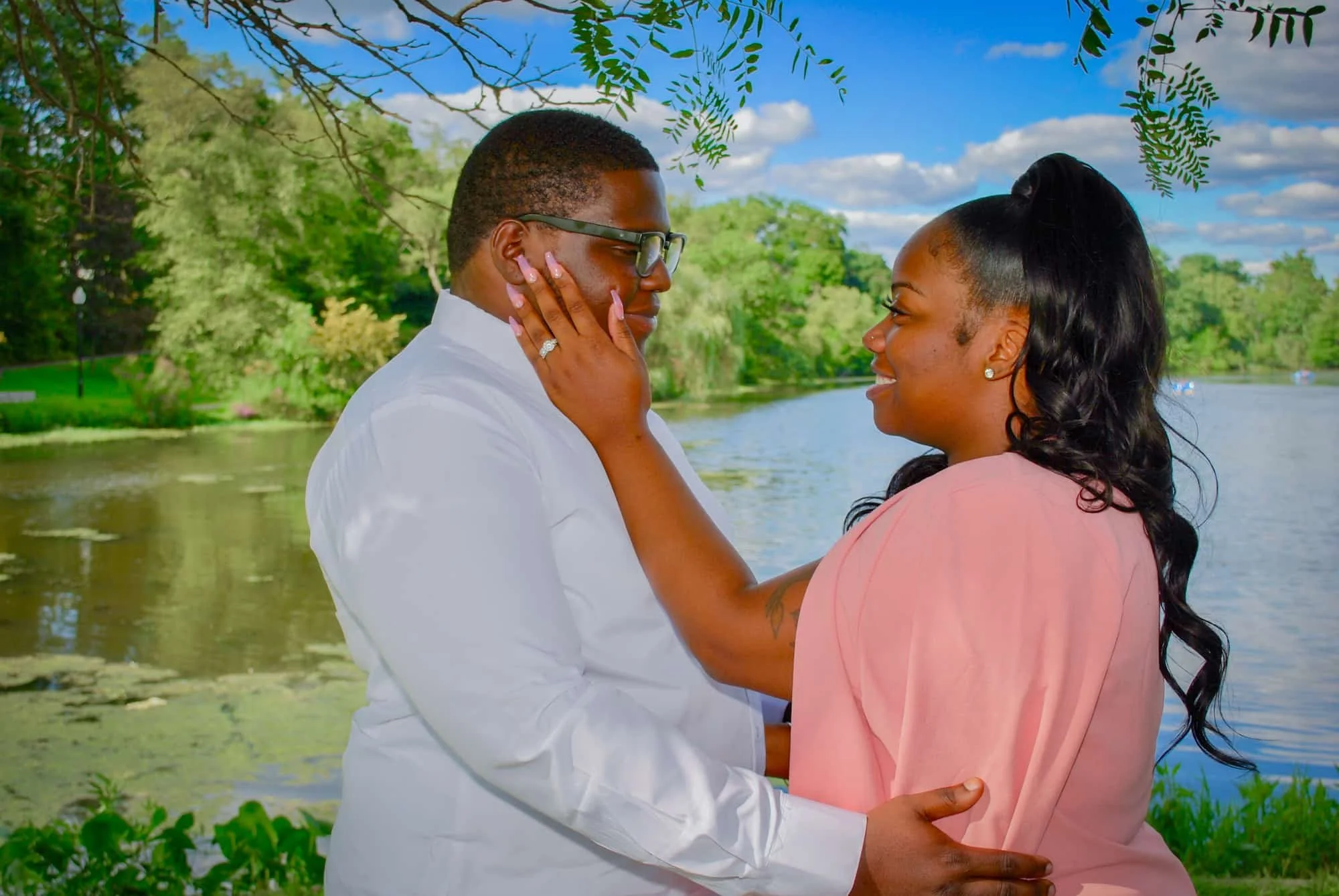 A couple gazing at each other by a river with trees and blue sky in the background, the woman gently touching the man's face.