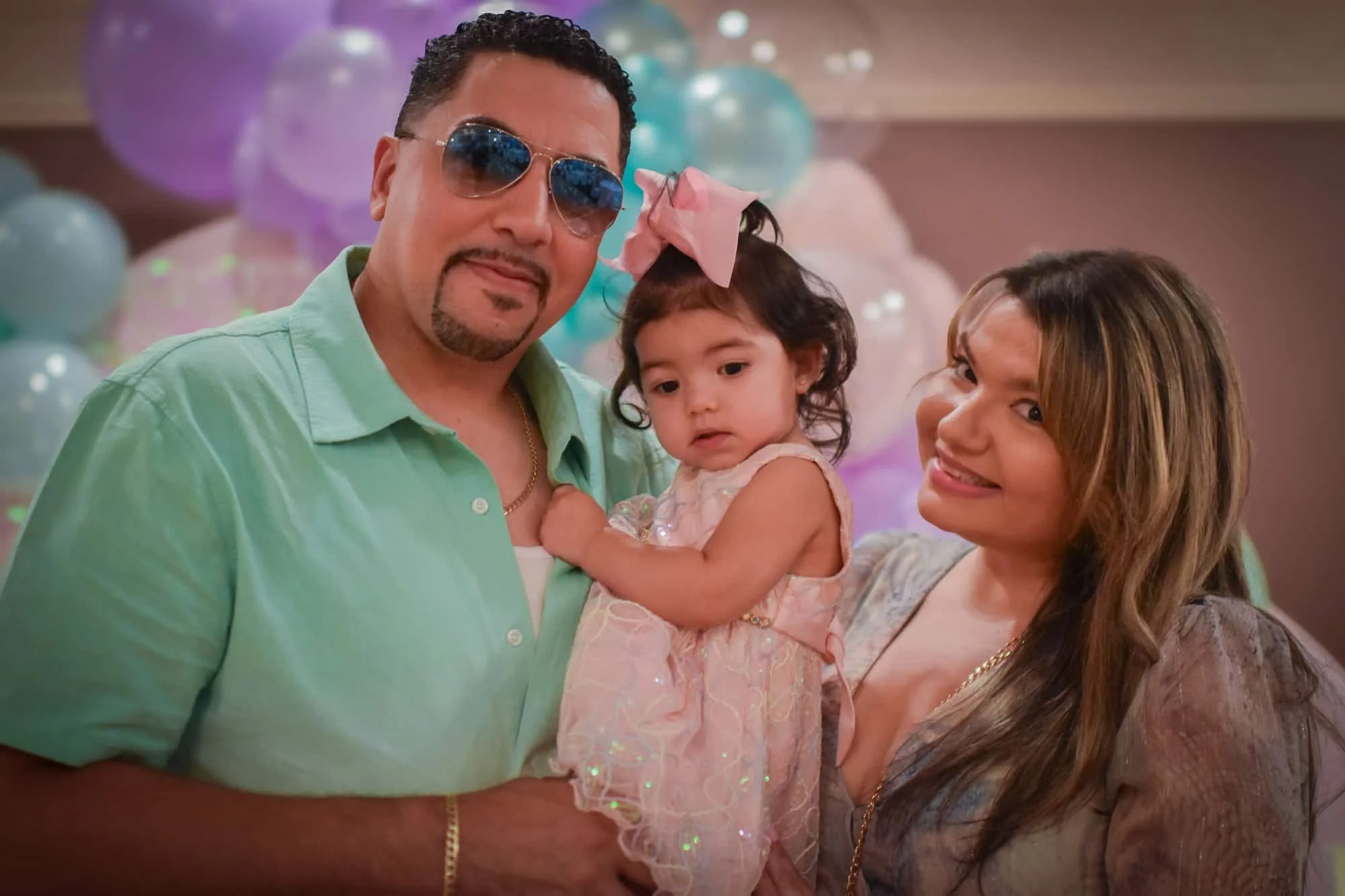 A man with sunglasses, a woman, and a young girl at a celebration with balloons in the background