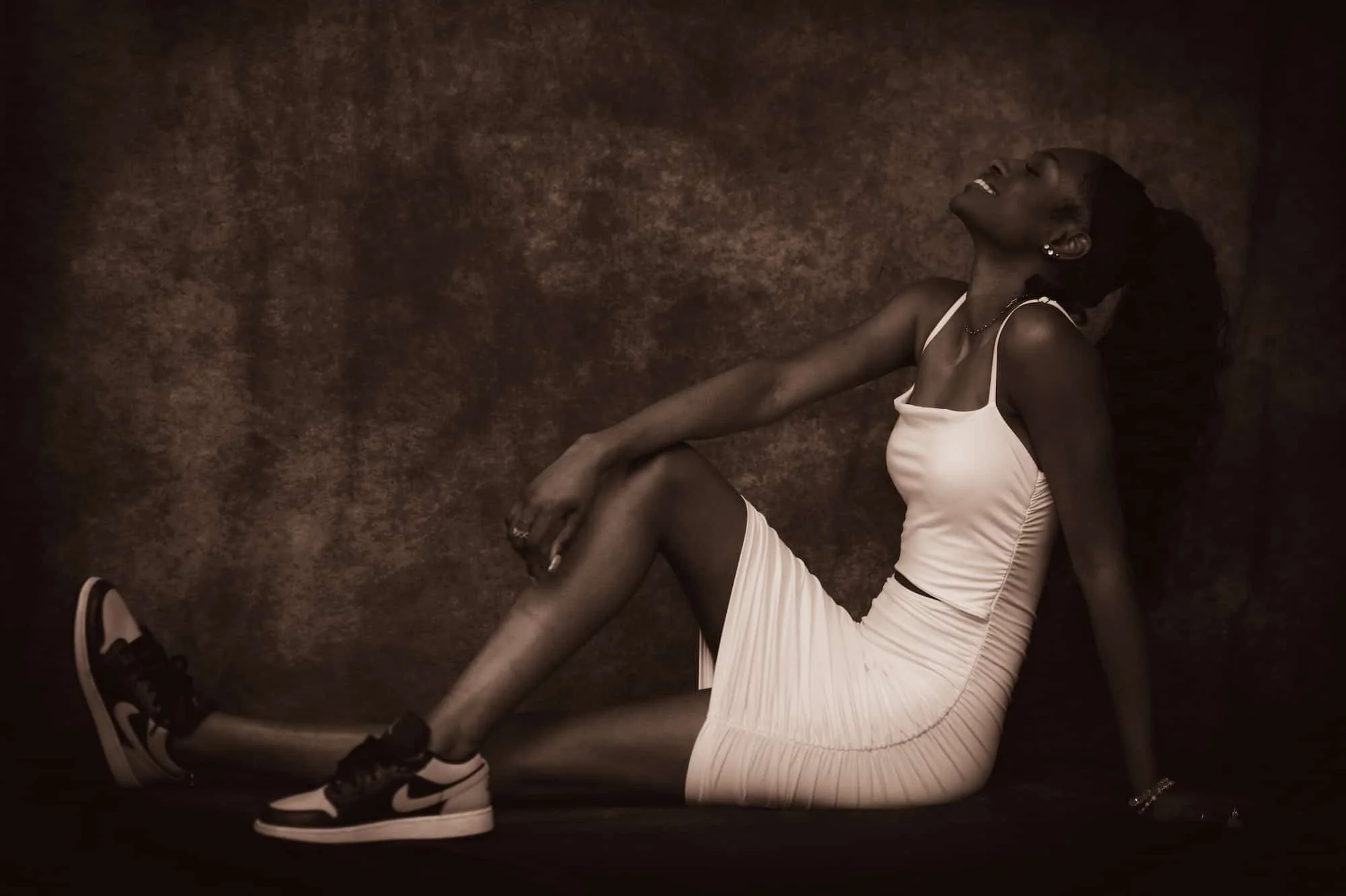 A young woman with dark skin wearing a white sleeveless dress and athletic sneakers, sitting on the floor with her legs extended and laughing with her head tilted back.