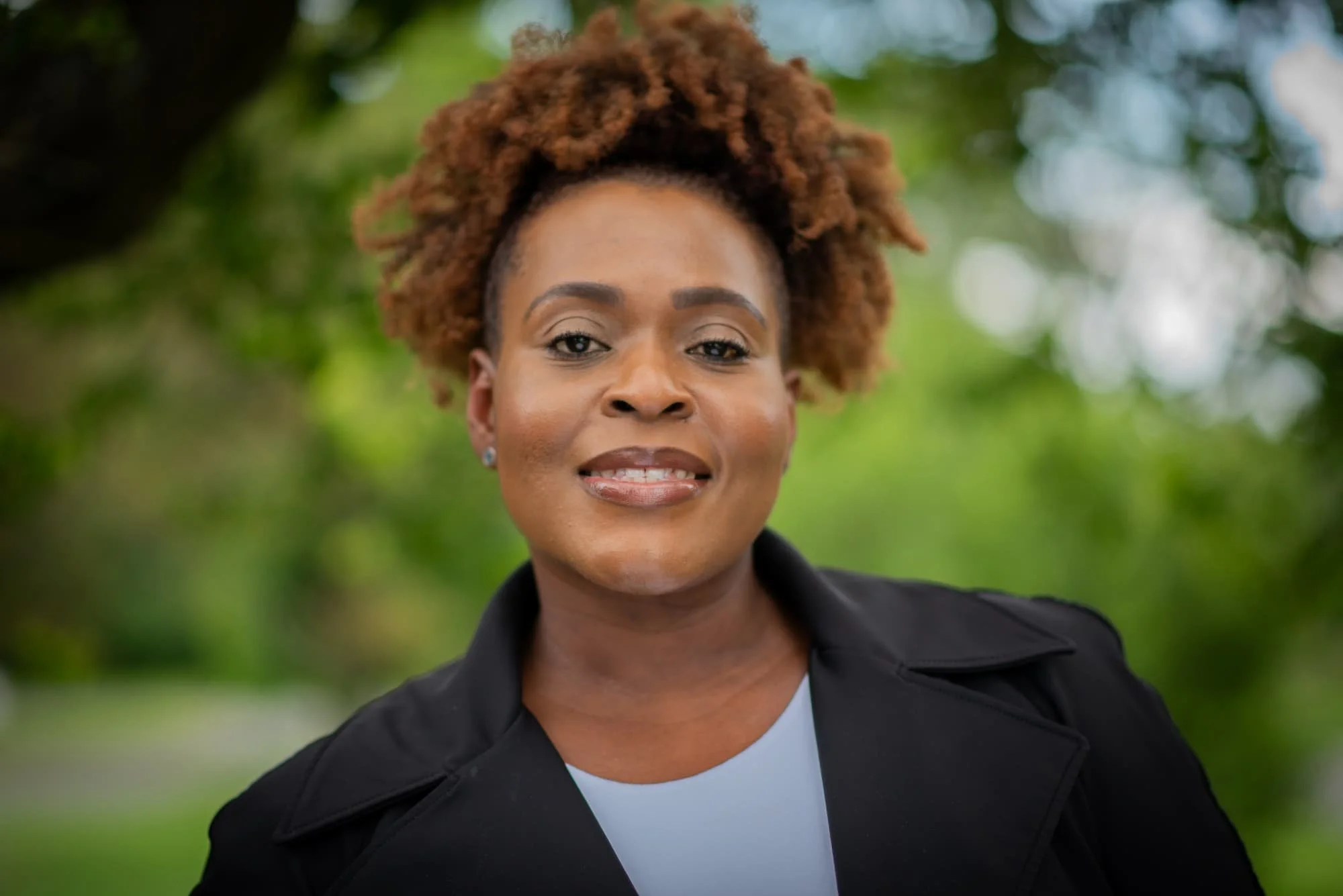A young woman with curly black hair, wearing glasses, hoop earrings, and a striped shirt, smiling indoors.