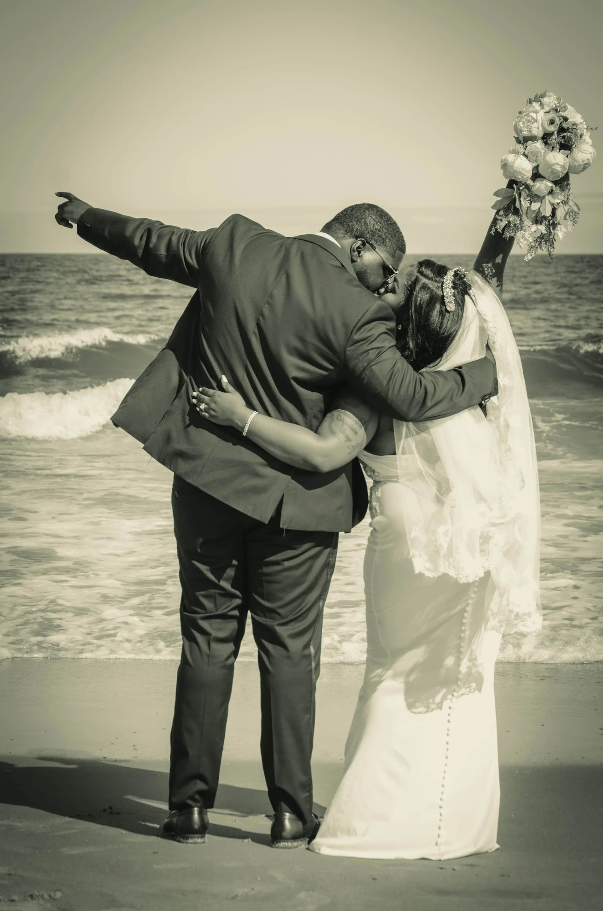 A couple in wedding attire sharing a kiss on the beach, with the ocean in the background and the bride holding a bouquet of flowers.