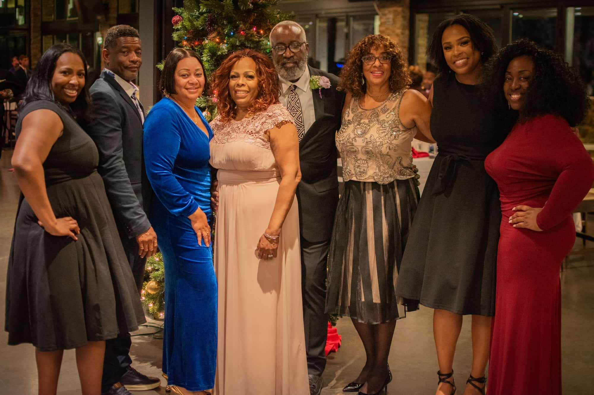 Group of nine people standing together near a decorated Christmas tree indoors, dressed in formal attire, smiling for the photo.