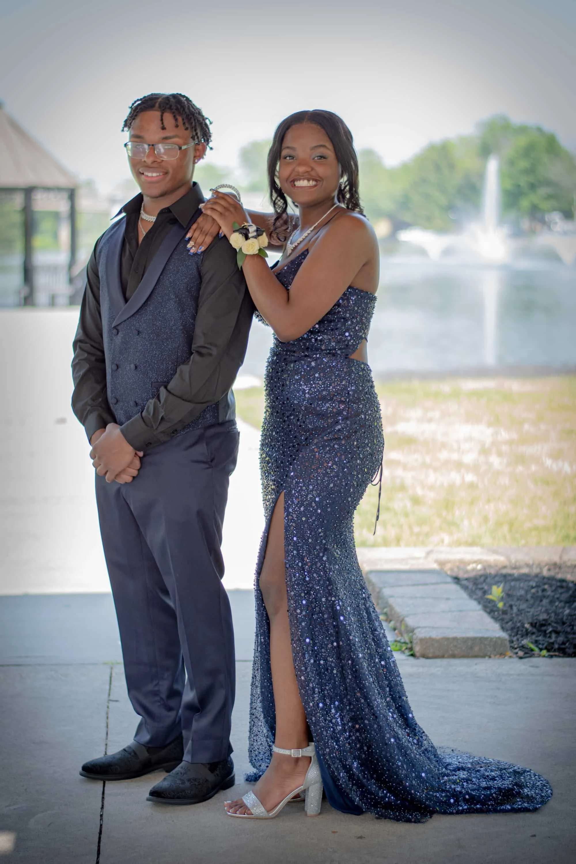 A young couple dressed in formal attire, standing outdoors near a body of water with fountains in the background. The girl wears a sparkly navy blue dress with a slit, and high-heeled sandals. The boy wears a dark suit with a vest and glasses. They a