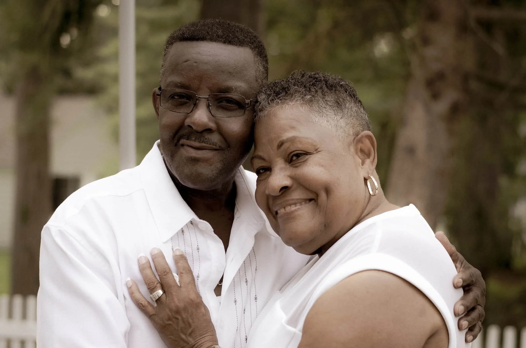 A middle-aged couple hugging outdoors, both smiling and wearing white shirts.