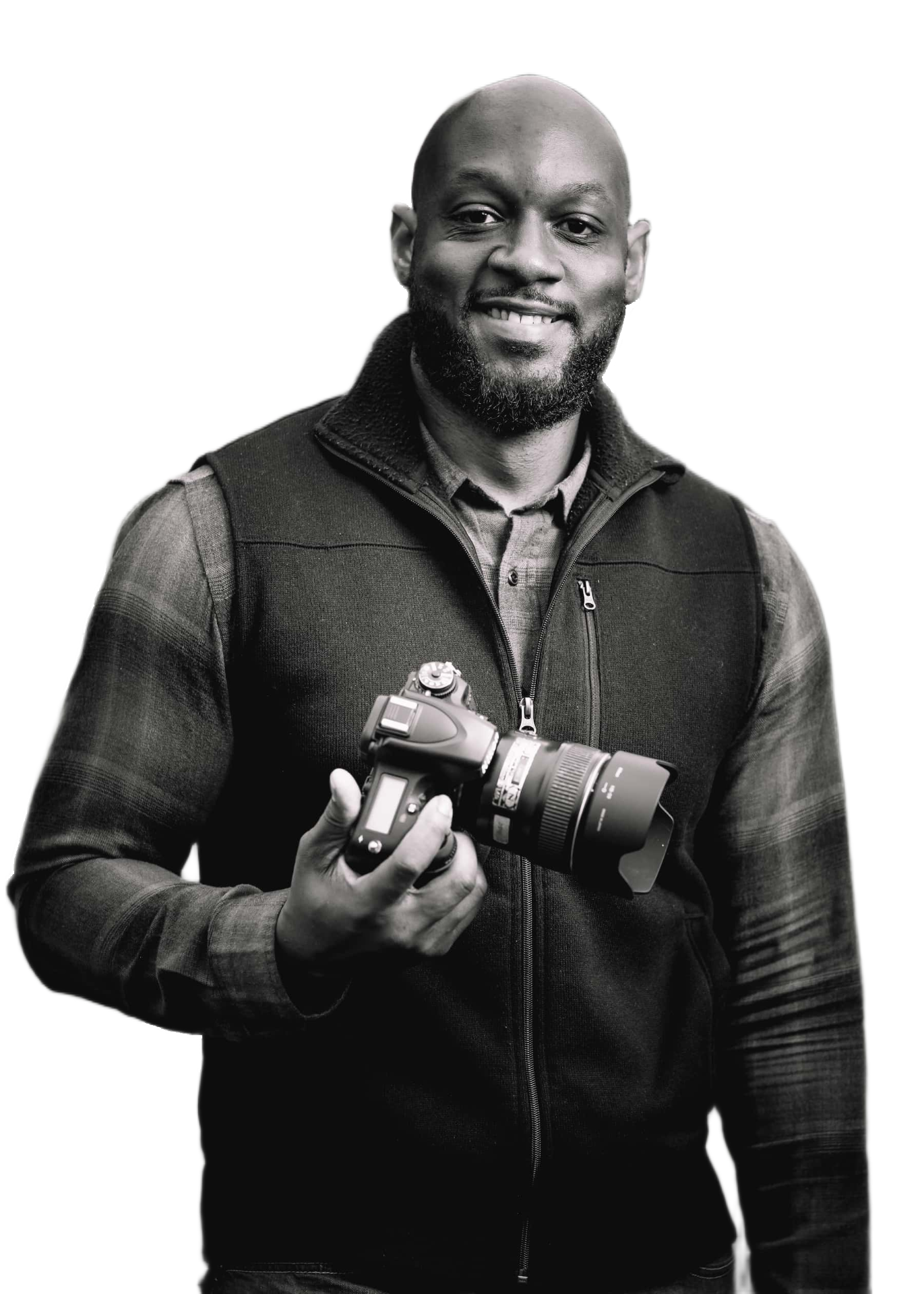 A smiling man holding a professional camera, wearing a dark vest over a checkered shirt, in black and white.