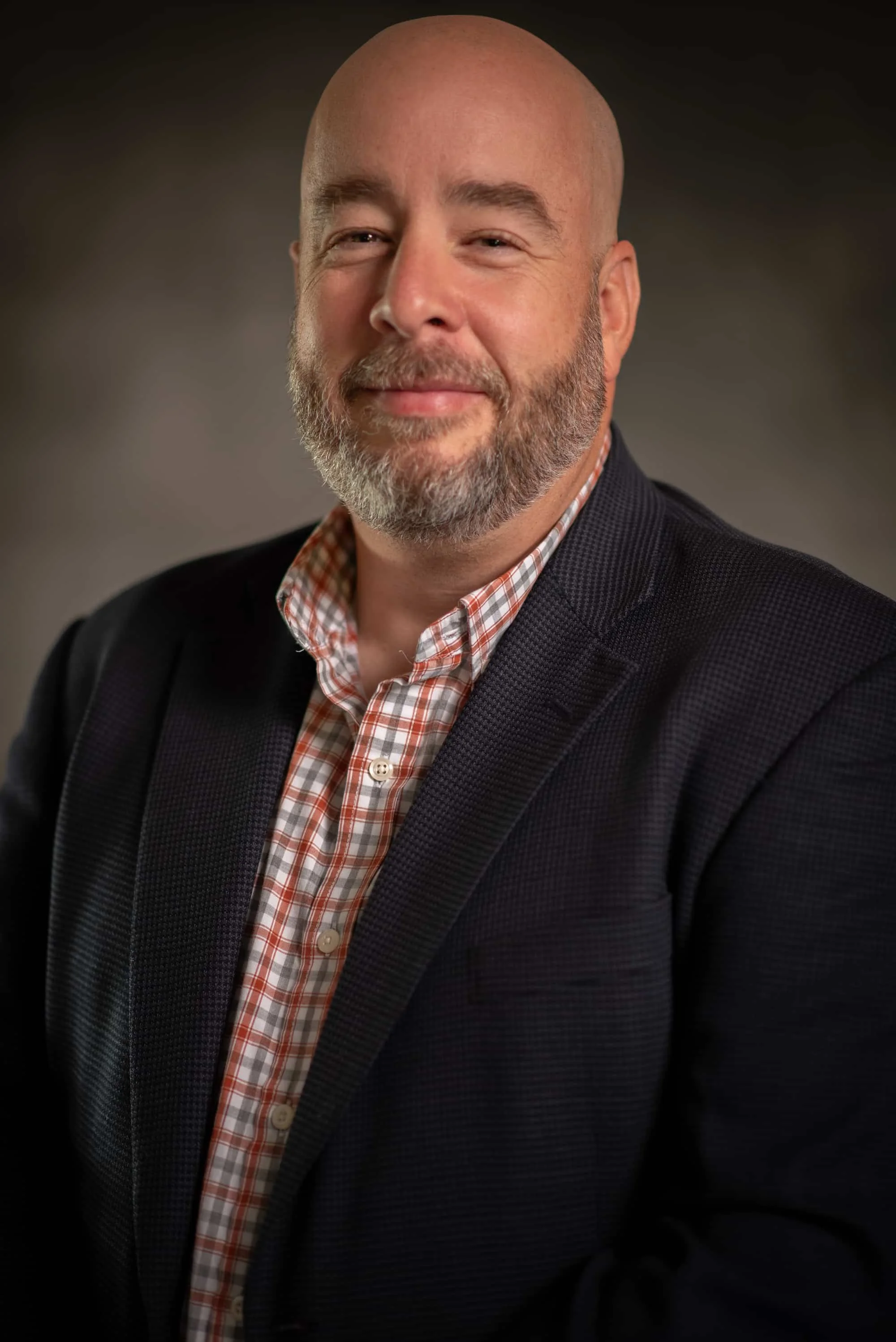 A middle-aged man with a beard, smiling, wearing a black blazer and a checkered button-up shirt, against a dark blurred background.