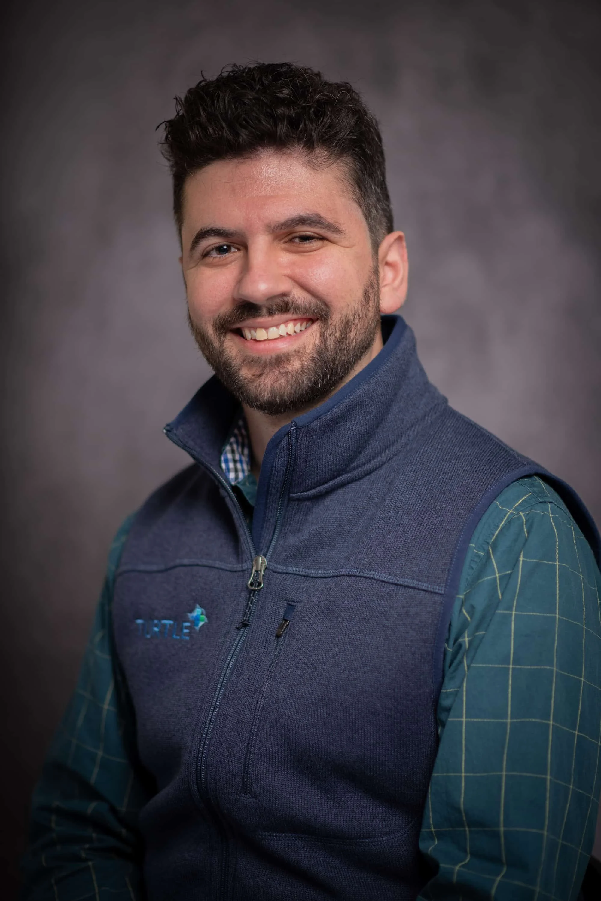 A man with dark, curly hair and a beard, smiling, wearing a blue fleece vest over a green plaid shirt, against a gray background.