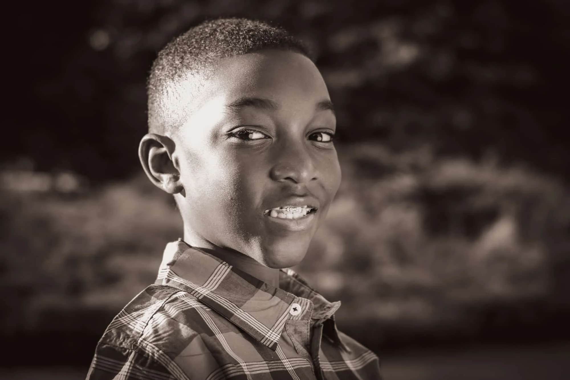 A young boy with short hair smiling in a plaid shirt, looking over his shoulder outdoors.