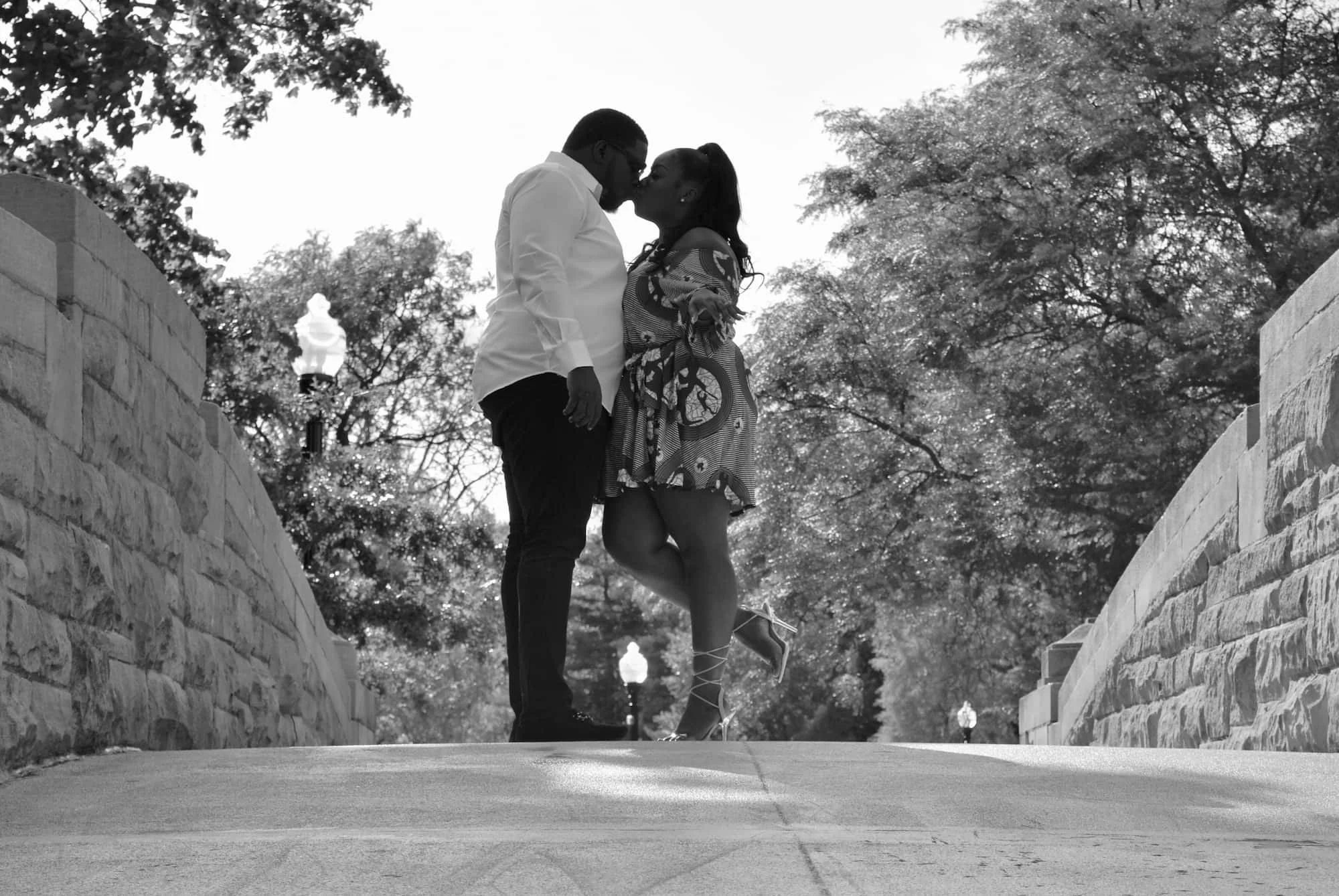 A black-and-white photo of a couple kissing on a bridge, with the man wearing a white shirt and the woman wearing a patterned dress and high heels, surrounded by trees and vintage-style street lamps.