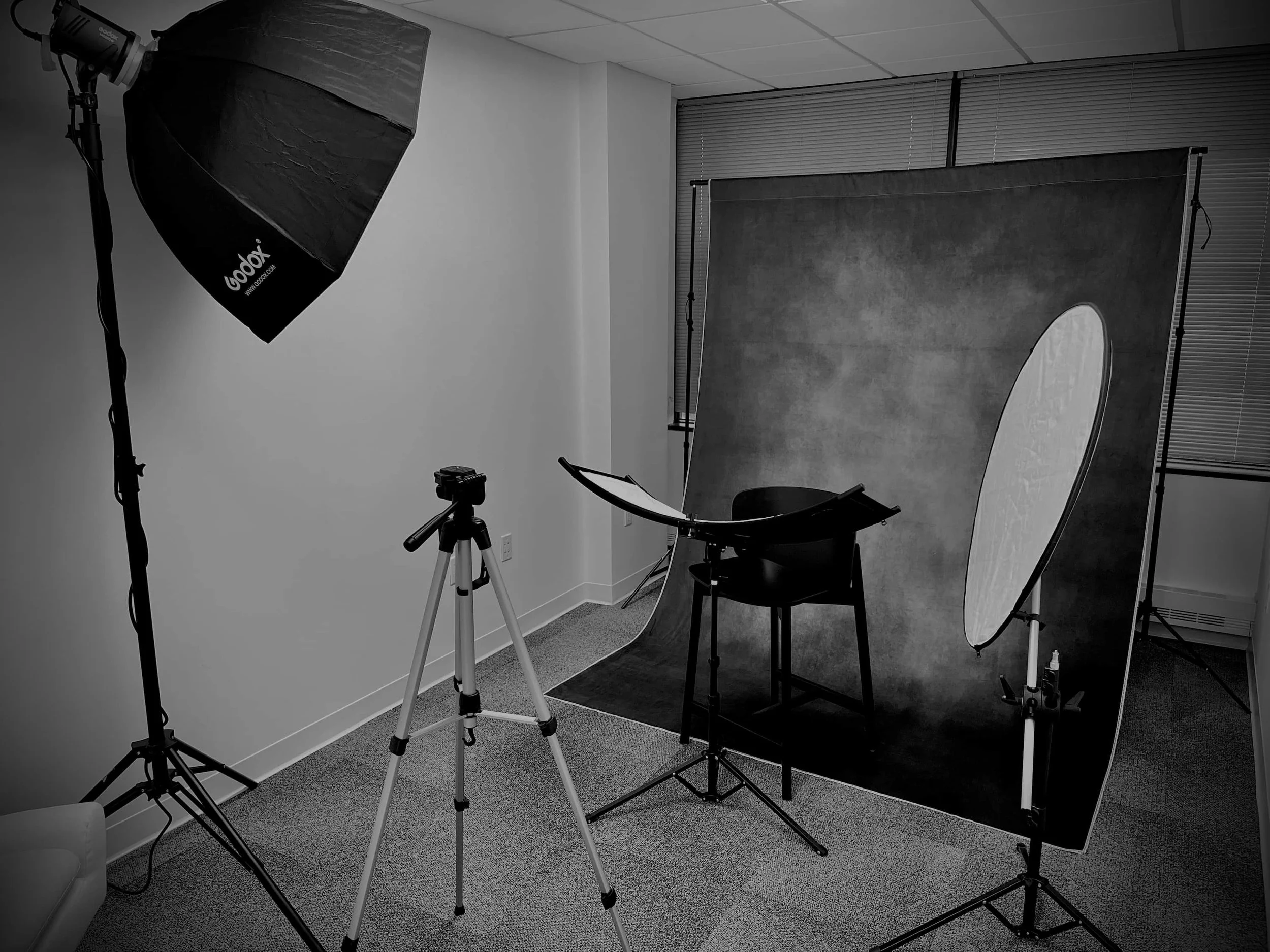 Photography studio setup with a backdrop, a chair, a professional light, a tripod, and photographic equipment in a room with window blinds.