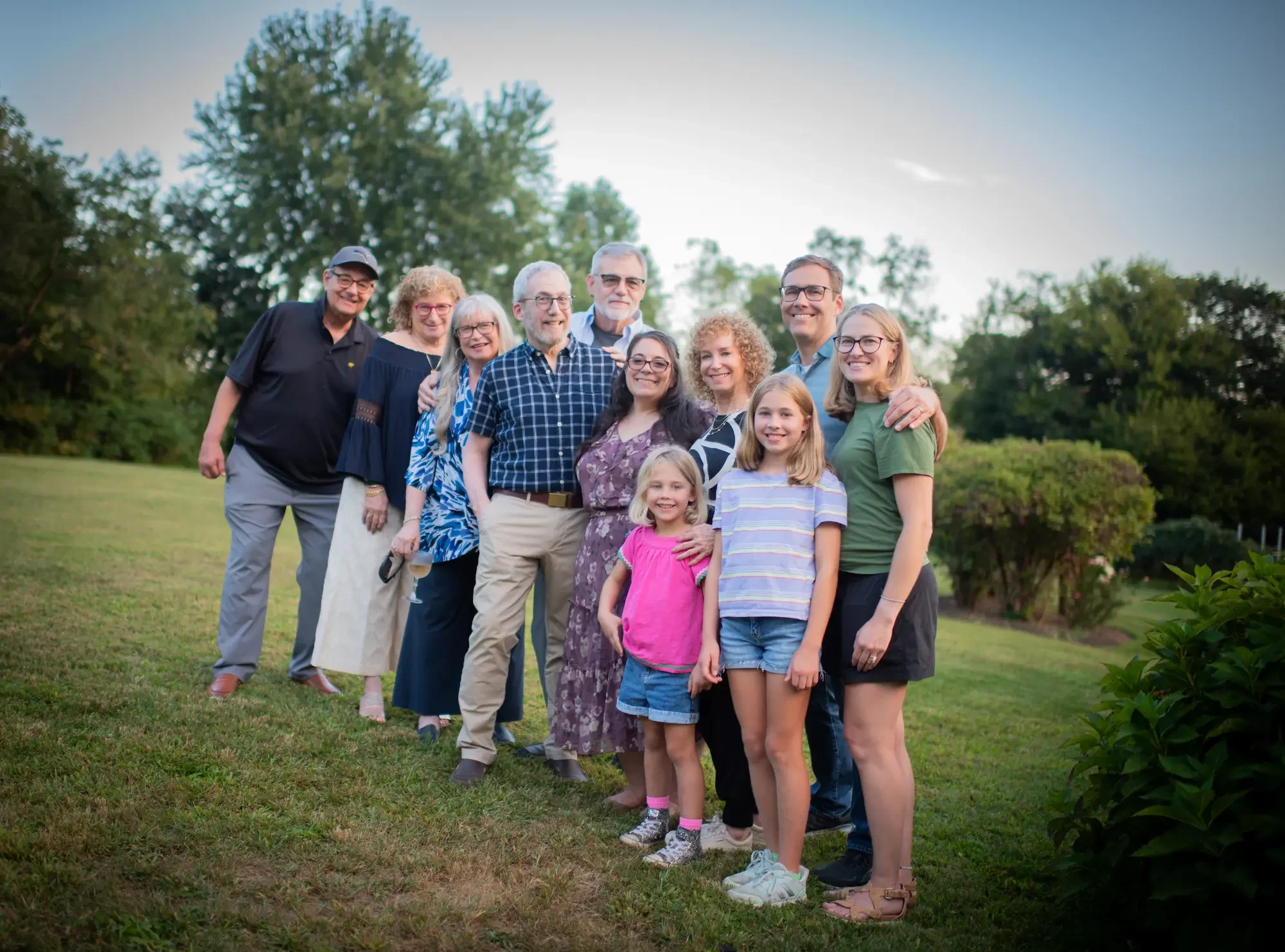 A multi-generational family group photo outdoors on a grassy field with trees in the background, taken during sunset or late afternoon.