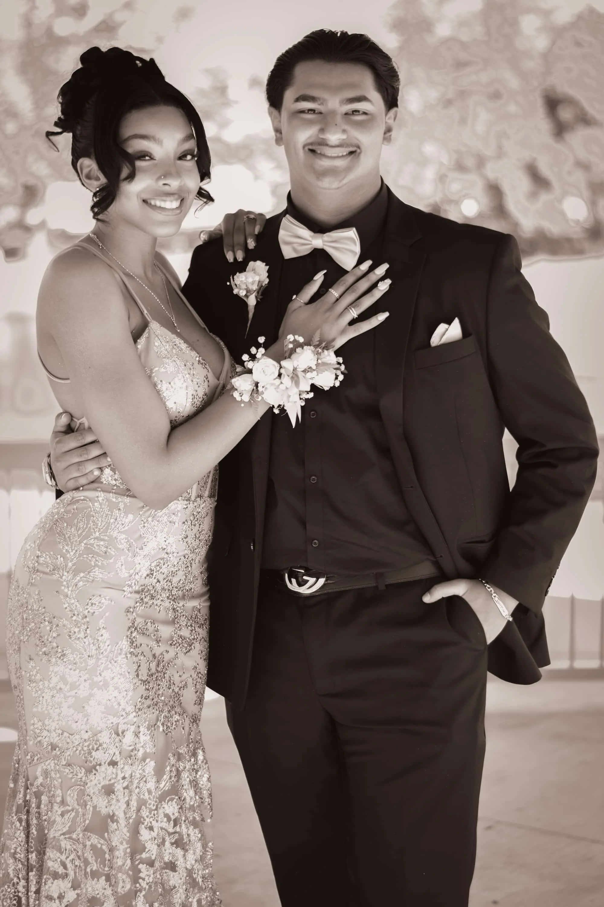 A couple in wedding attire smiling and posing for a photo indoors, with the woman wearing a lace wedding dress and the man in a dark suit with a bow tie.