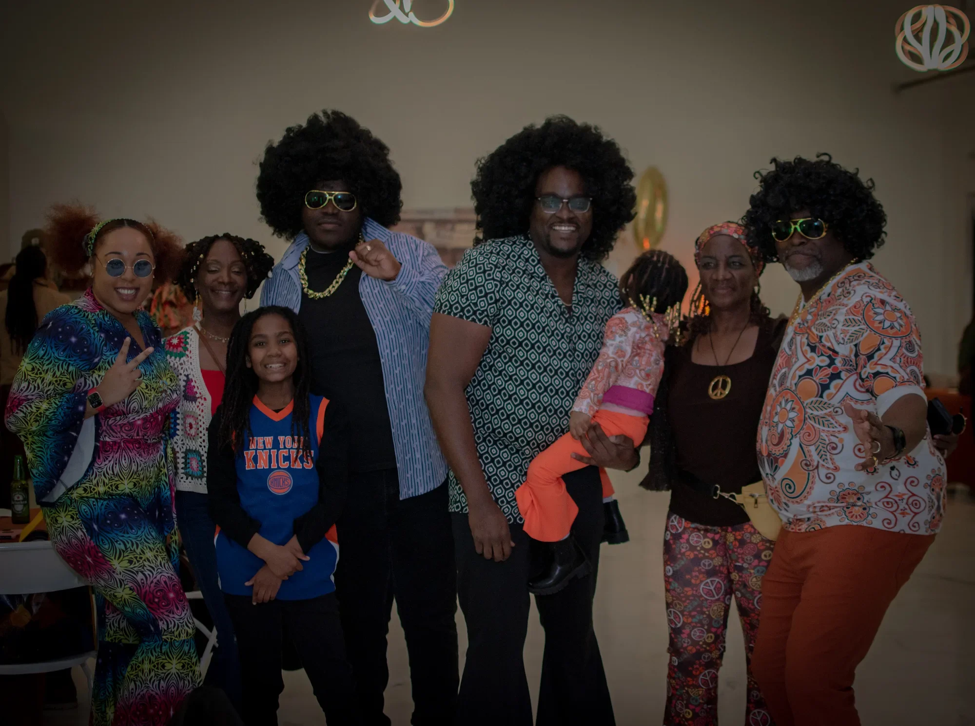 A group of people dressed in colorful, 70s-inspired outfits, some wearing Afro wigs and sunglasses, posing together at a party with peace sign accessories, with a mix of women, a man, and a child.