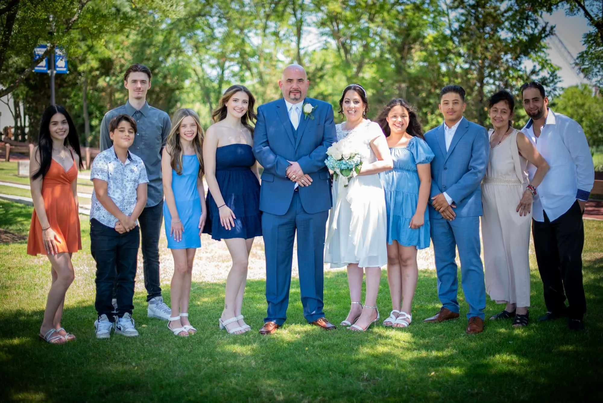 A group of people dressed for a wedding, standing outdoors on grass with trees in the background. The bride and groom are in the center, with the bride holding a bouquet of white flowers. They are surrounded by family and friends in colorful dresses 