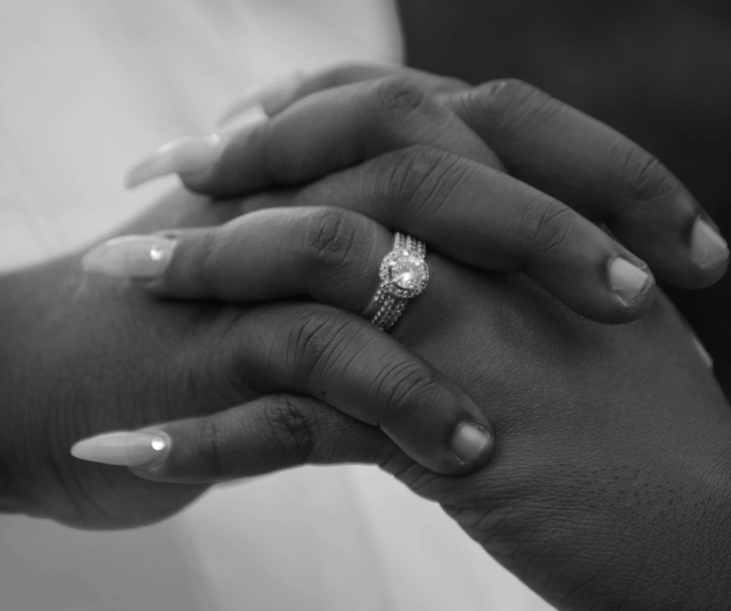 Two hands with intertwined fingers, one wearing a diamond engagement ring and a wedding band, in black and white.