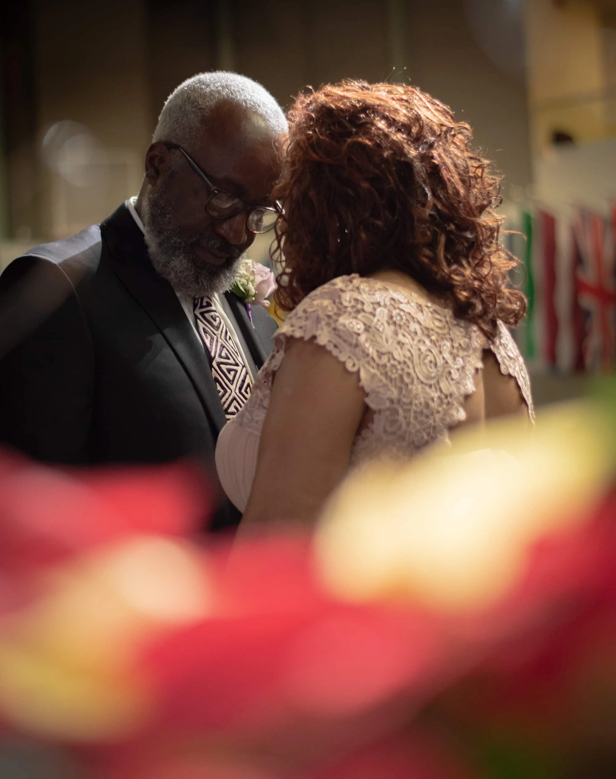 A couple during their wedding ceremony, with the groom wearing glasses and a suit, and the bride with curly hair in a lace dress, standing close together.