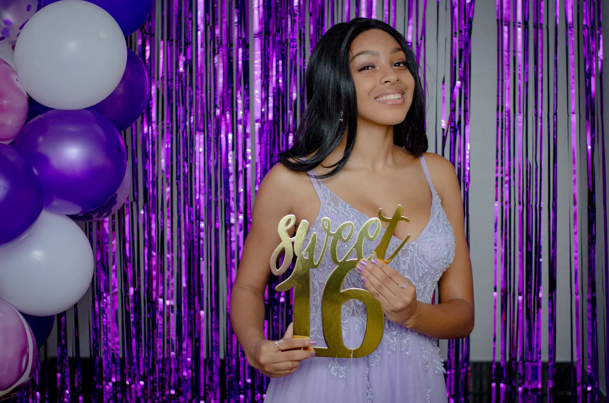 A woman at her 16th birthday celebration standing in front of a purple and black metallic fringe backdrop, holding a gold 'sweet 16' sign, smiling and wearing a lavender dress.
