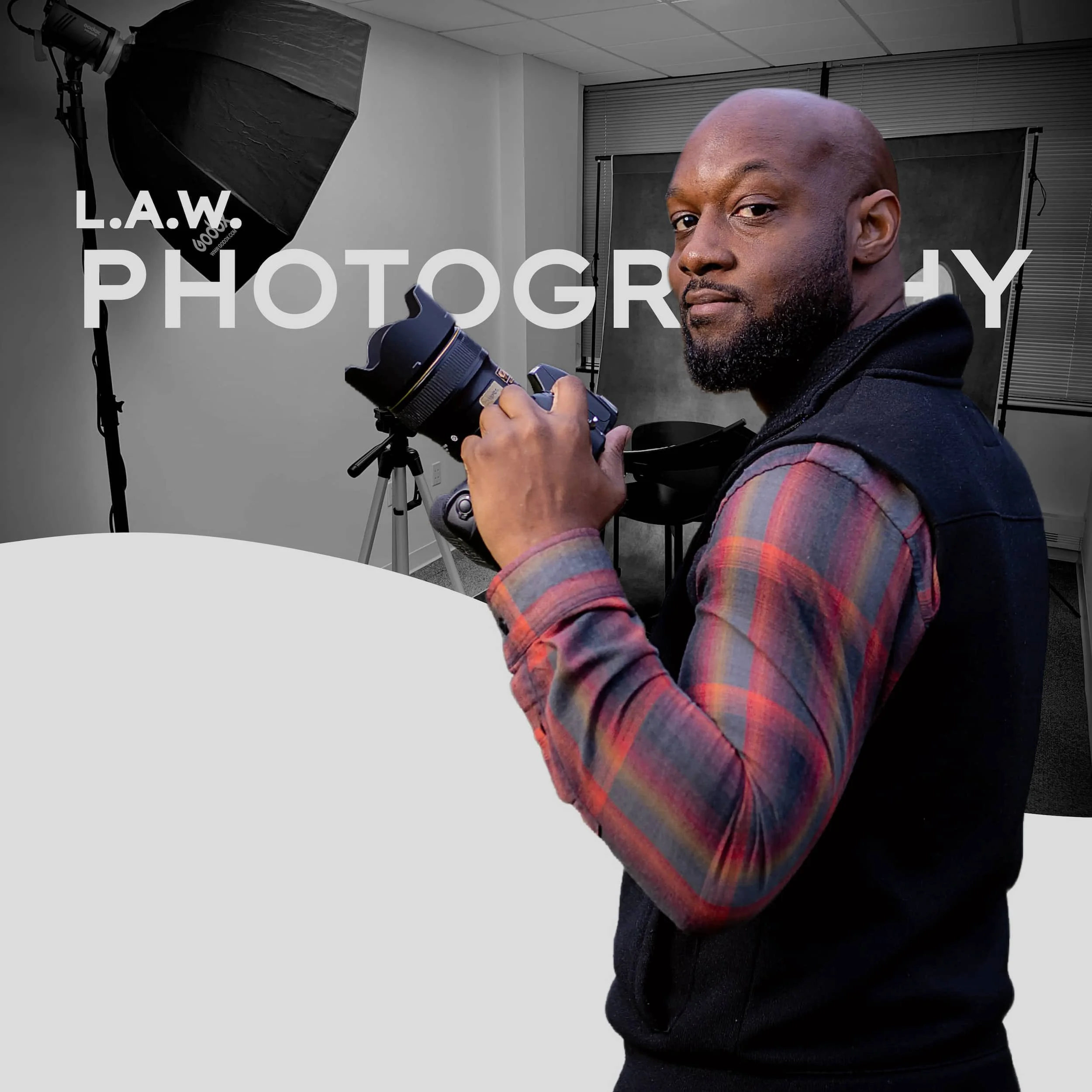 A man holding a professional camera, in a photography studio with lighting equipment, gray backdrop, and furniture.
