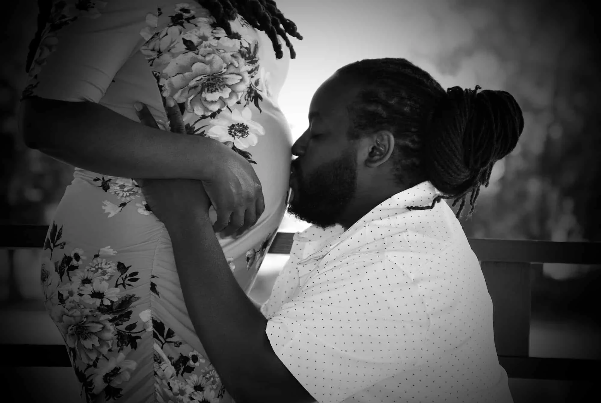 A man kisses a pregnant woman on her belly while holding her waist.