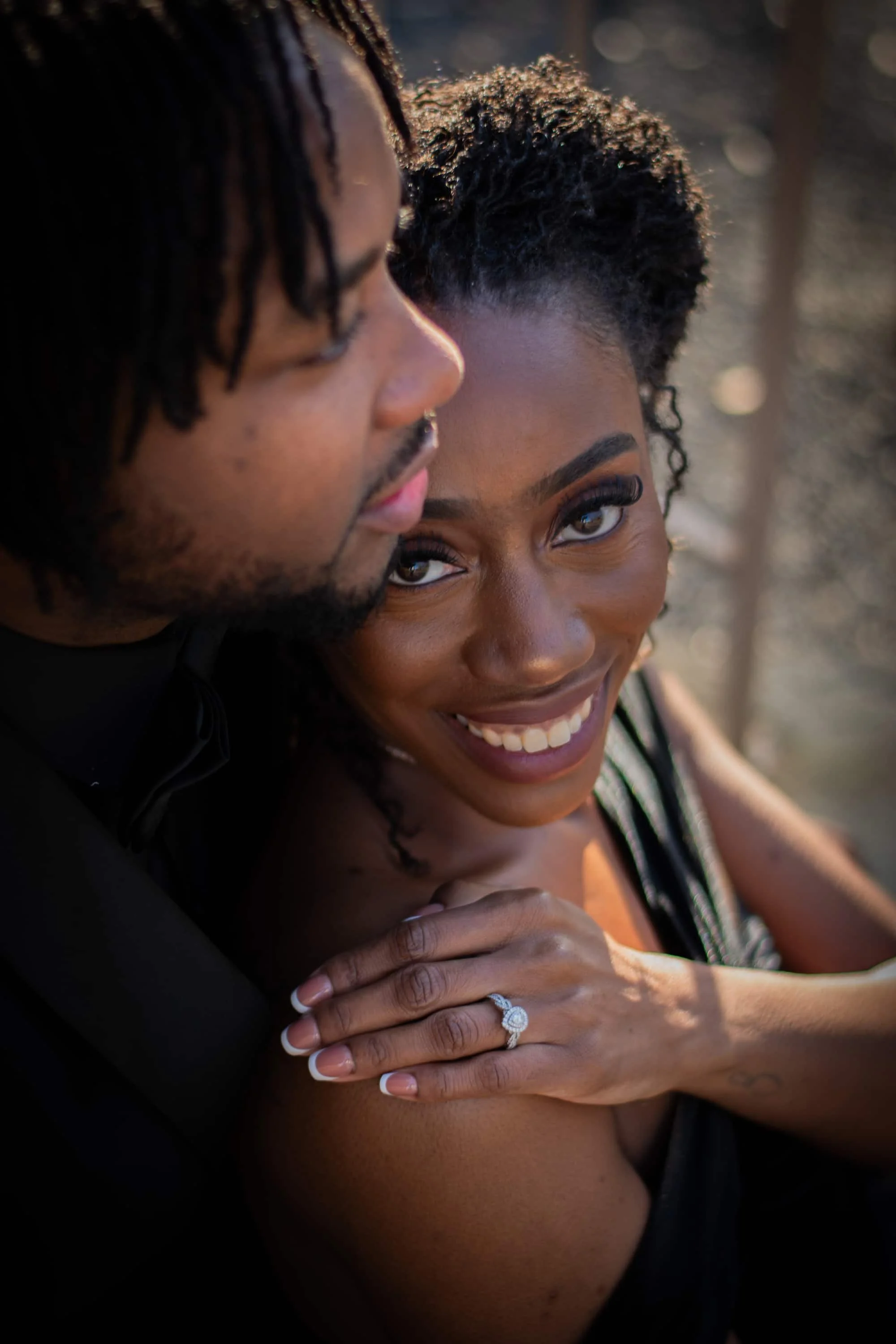 A smiling black woman showing off an engagement ring on her finger while leaning on a man with dreadlocks, both looking happy and intimate.